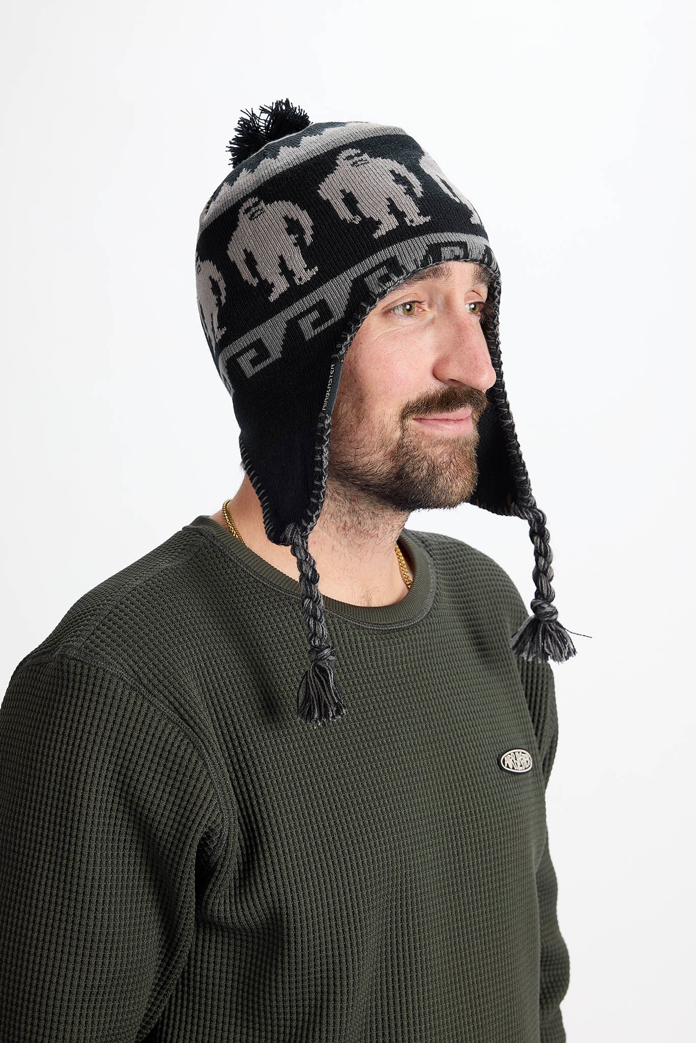 A man wearing a dark green waffle-knit crewneck sweatshirt and a black knit beanie with gray silhouettes and ear flaps, photographed against a plain white background.