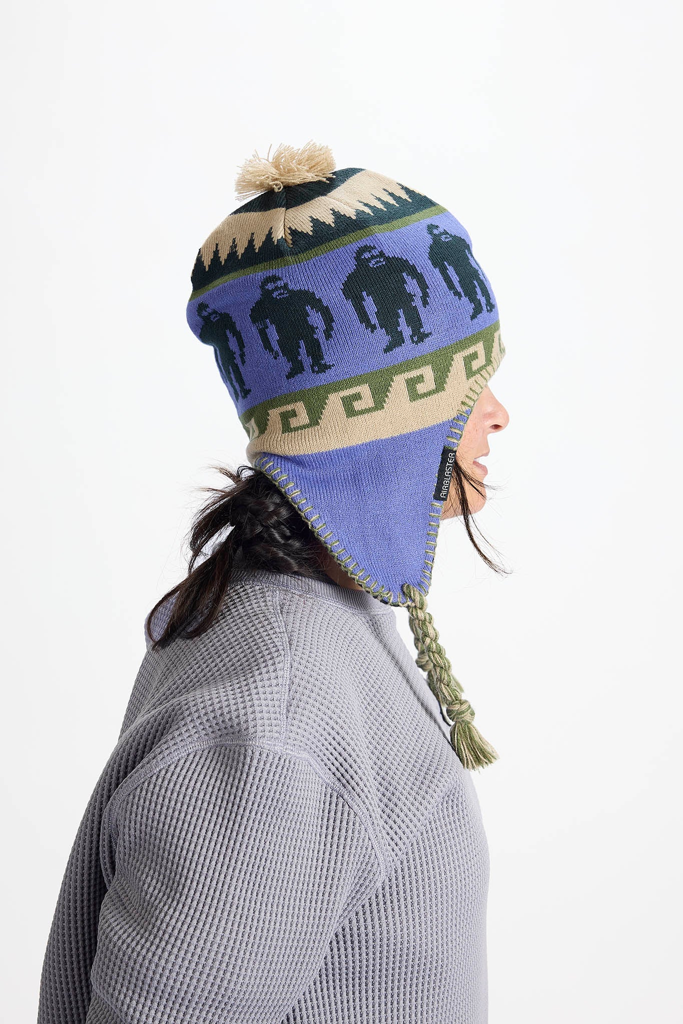 A side-profile view of a person wearing a knit winter hat with ear flaps and a pom-pom. The hat features a blue-purple base with black gorilla silhouettes, a beige geometric band, and a green patterned section near the brim.