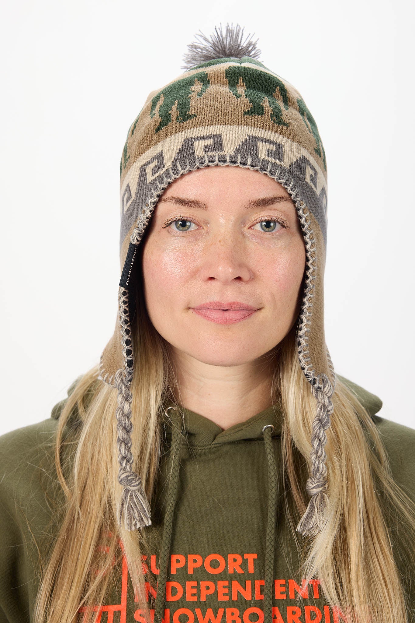 A woman wearing a camouflage-patterned knit winter hat with ear flaps and a pom-pom, paired with an olive green hoodie against a plain white background.