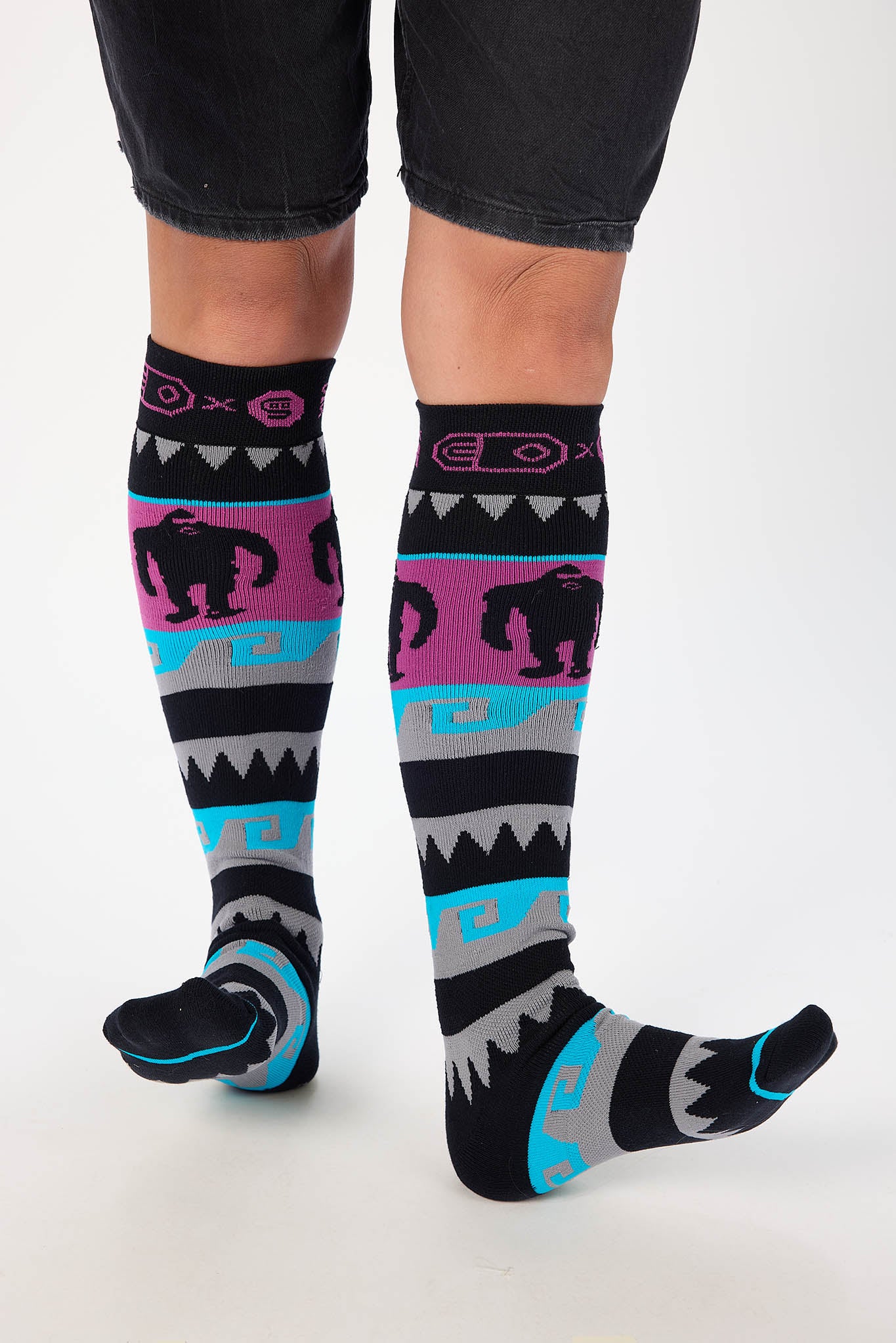 A person wearing black knee-high socks with a colorful Southwestern-style pattern featuring pink, turquoise, gray, and black geometric stripes and gorilla silhouettes, paired with black shorts against a white studio background.