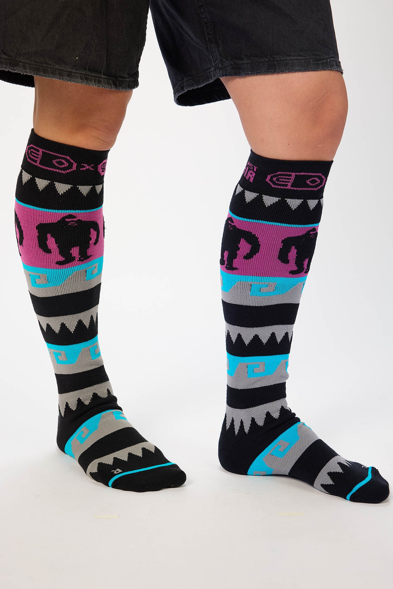 A person wearing knee-high patterned socks with black, pink, turquoise, and gray bands, shown from the calves down against a light studio background.