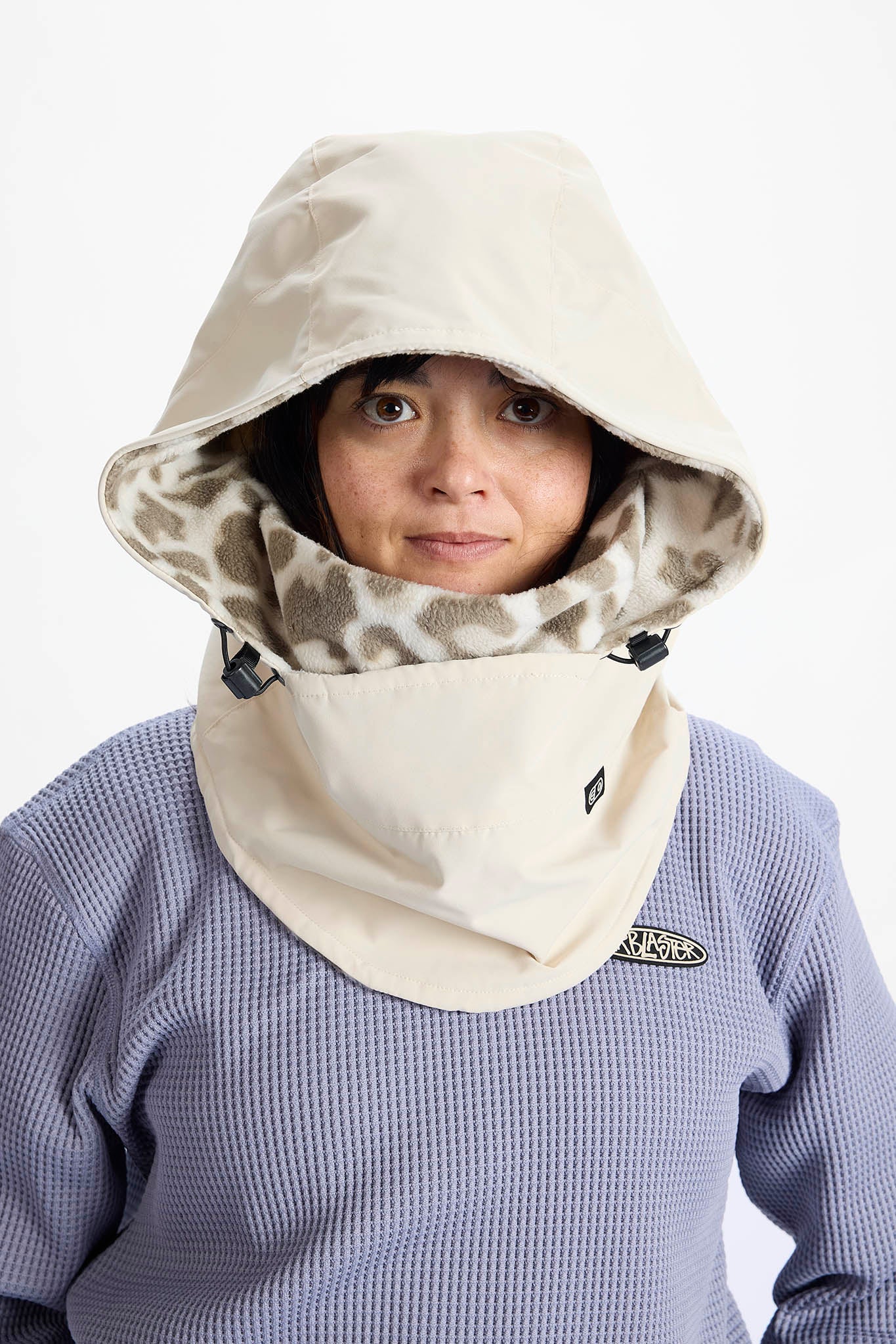 A person wearing a beige hooded neck warmer with a patterned inner lining and a waffle-knit lavender shirt underneath, against a plain white background.