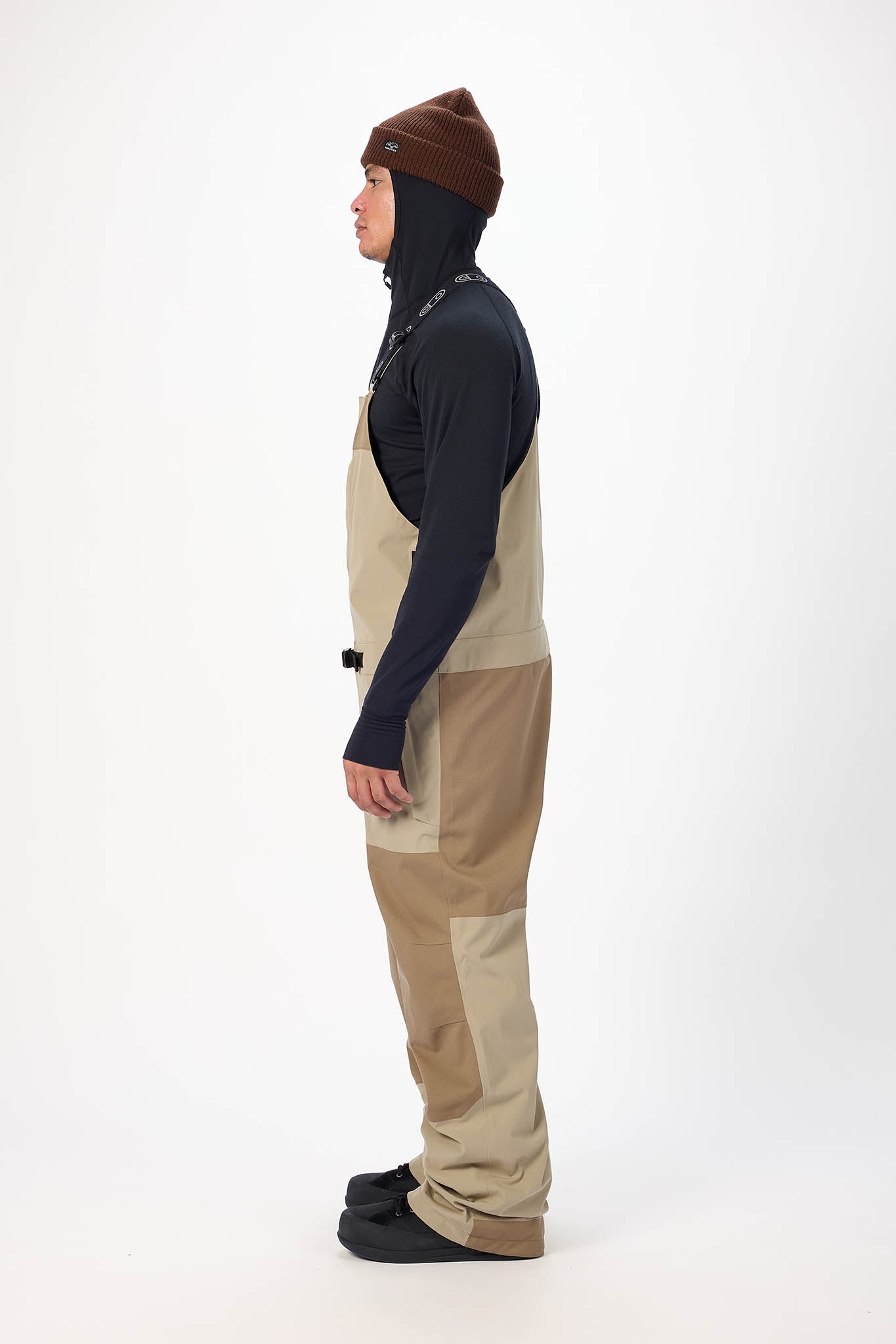 Person wearing a dark jacket, brown beanie, and beige pants on a white background