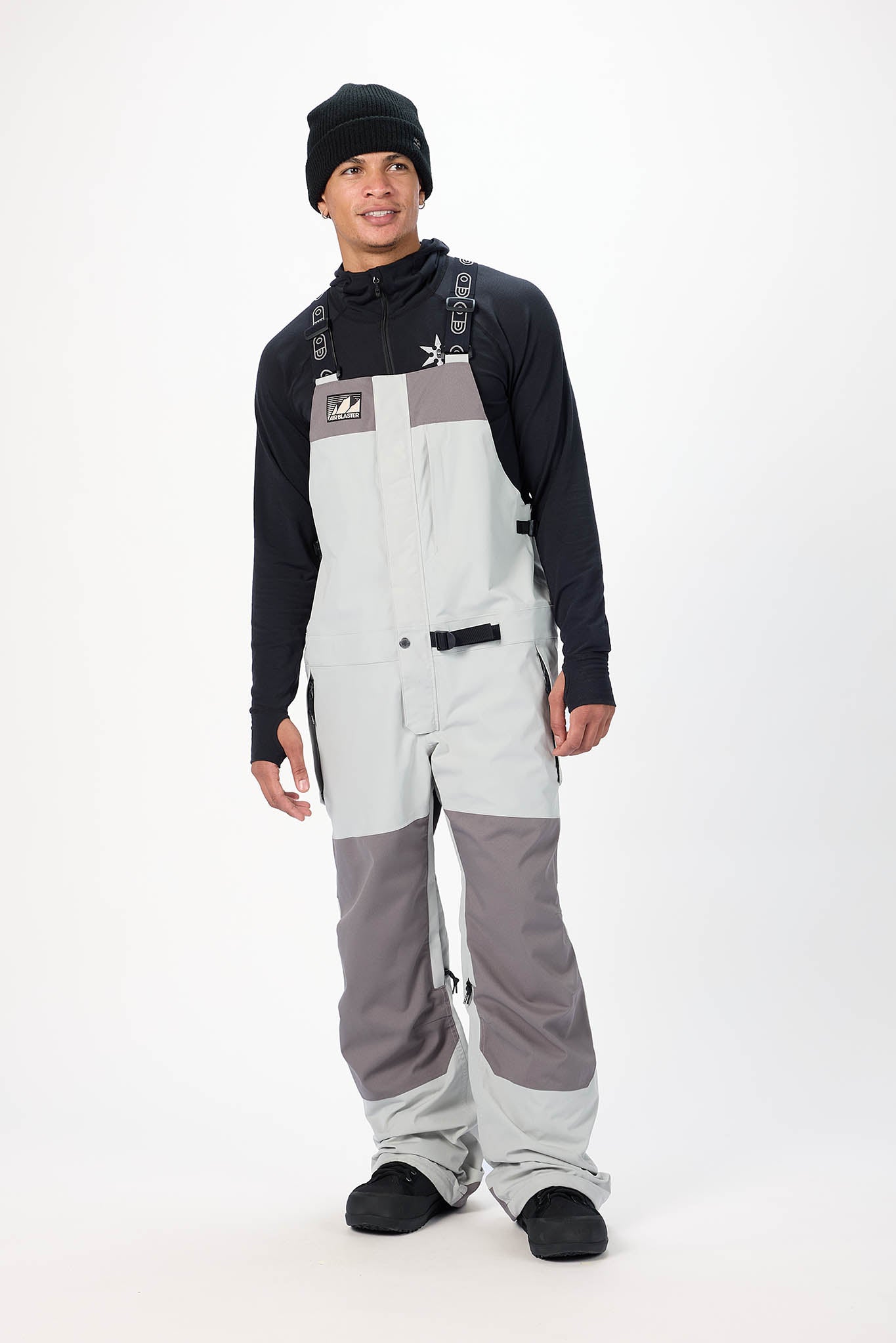 Person wearing a black jacket, gray bib overalls, and black boots on a white background