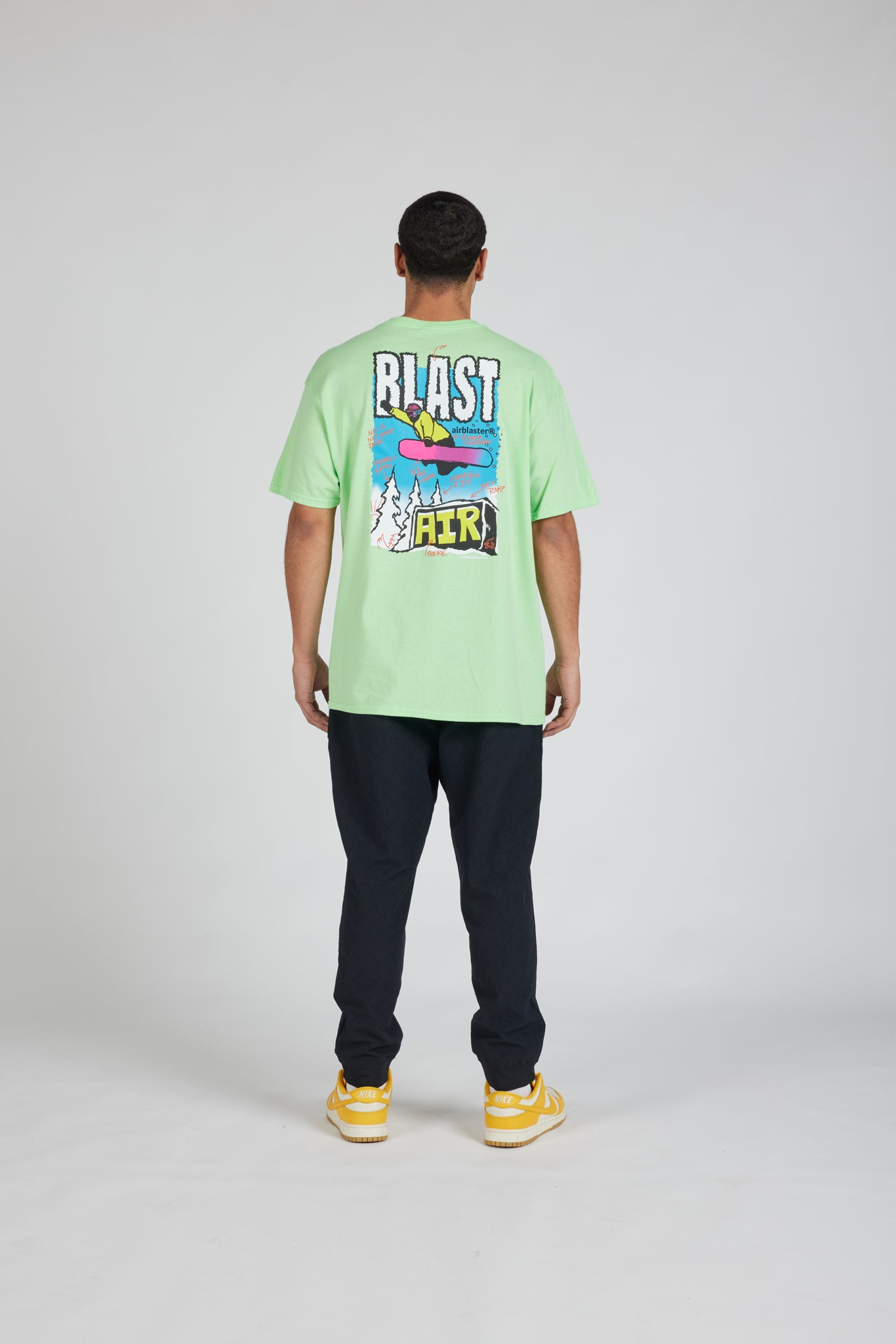 A man viewed from the back wearing a light green t-shirt with a colorful graphic print on the back, paired with dark pants and yellow sneakers, against a light gray studio background.