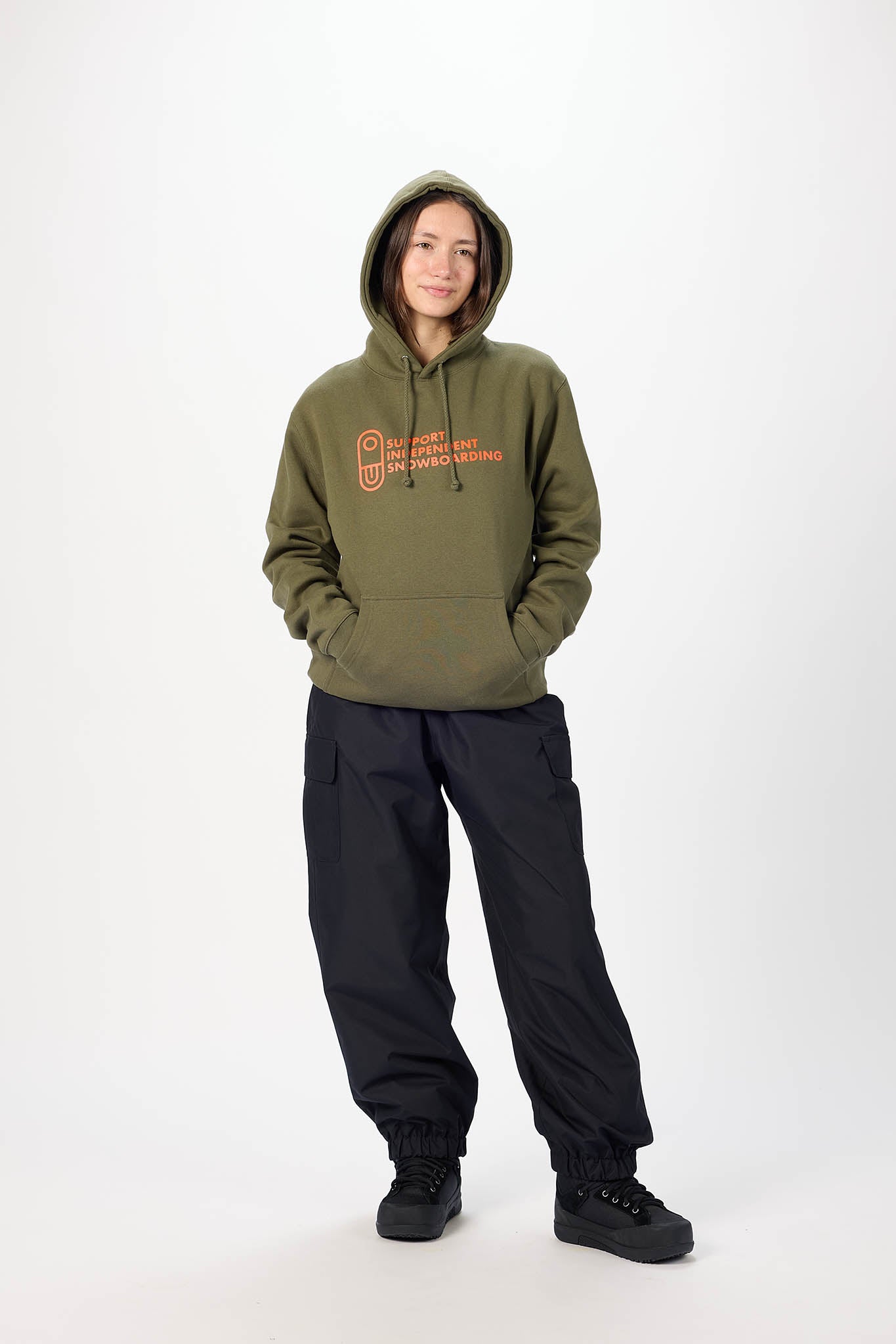 A model stands against a plain white studio background wearing an olive green pullover hoodie with a red and orange graphic on the chest, paired with black pants and black sneakers.