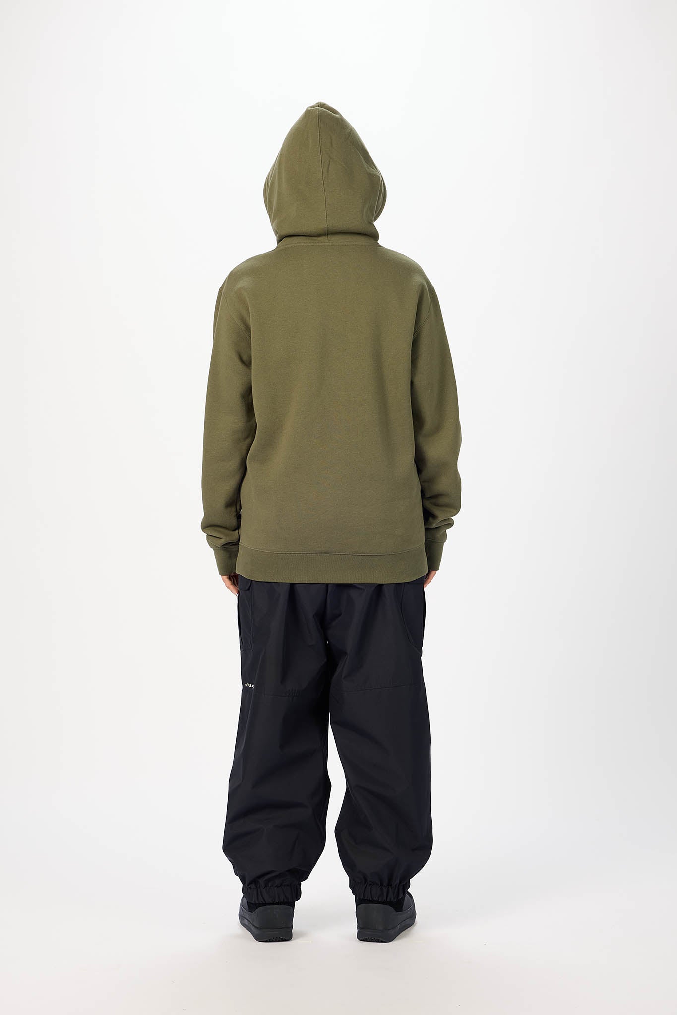 A person shown from the back wearing an olive green hooded sweatshirt and loose black pants, standing against a plain light gray studio background.