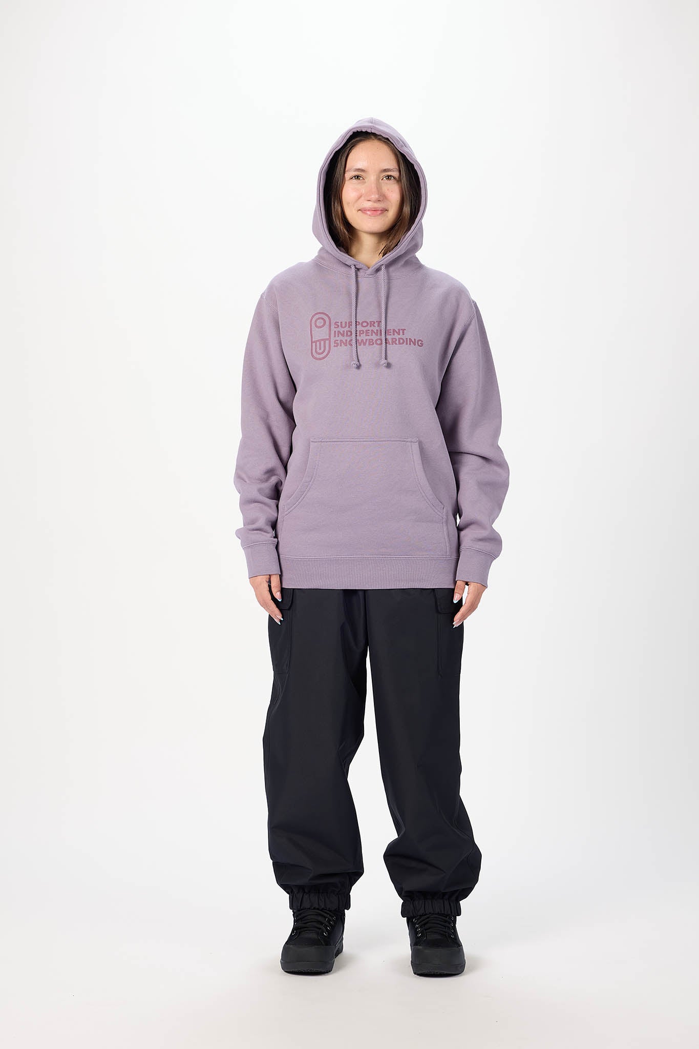 A model wearing a lavender-purple pullover hoodie with a pink graphic logo on the chest, paired with black sweatpants and black sneakers, standing against a plain white studio background.