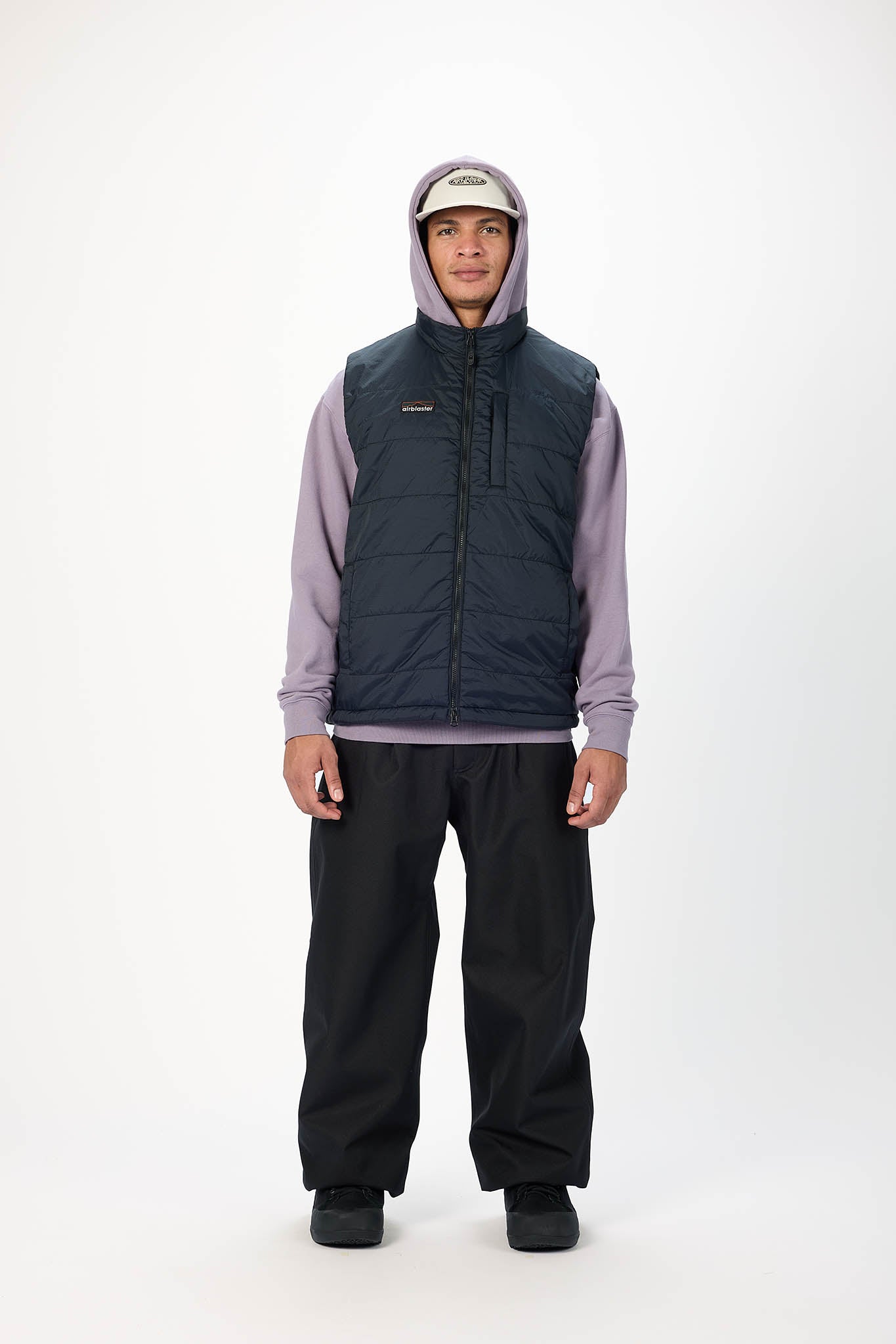 A model stands against a white studio background wearing a dark navy sleeveless puffer vest over a light purple hoodie, black pants, and a beige baseball cap.