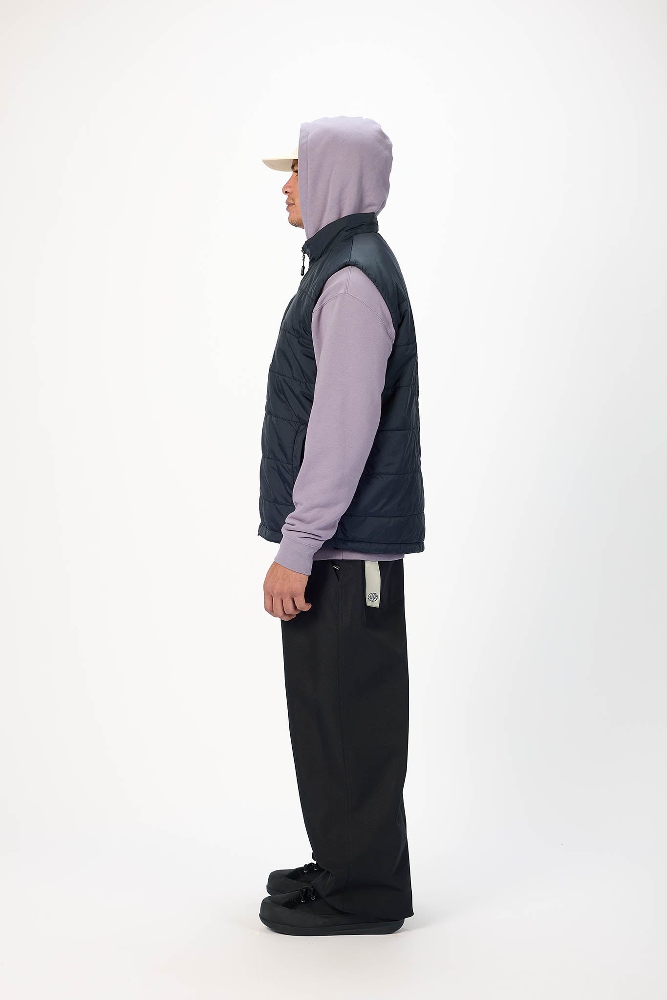 A person shown in profile wearing a dark navy sleeveless puffer vest over a light purple long-sleeve top, black pants, and a beige cap, standing against a plain white studio background.