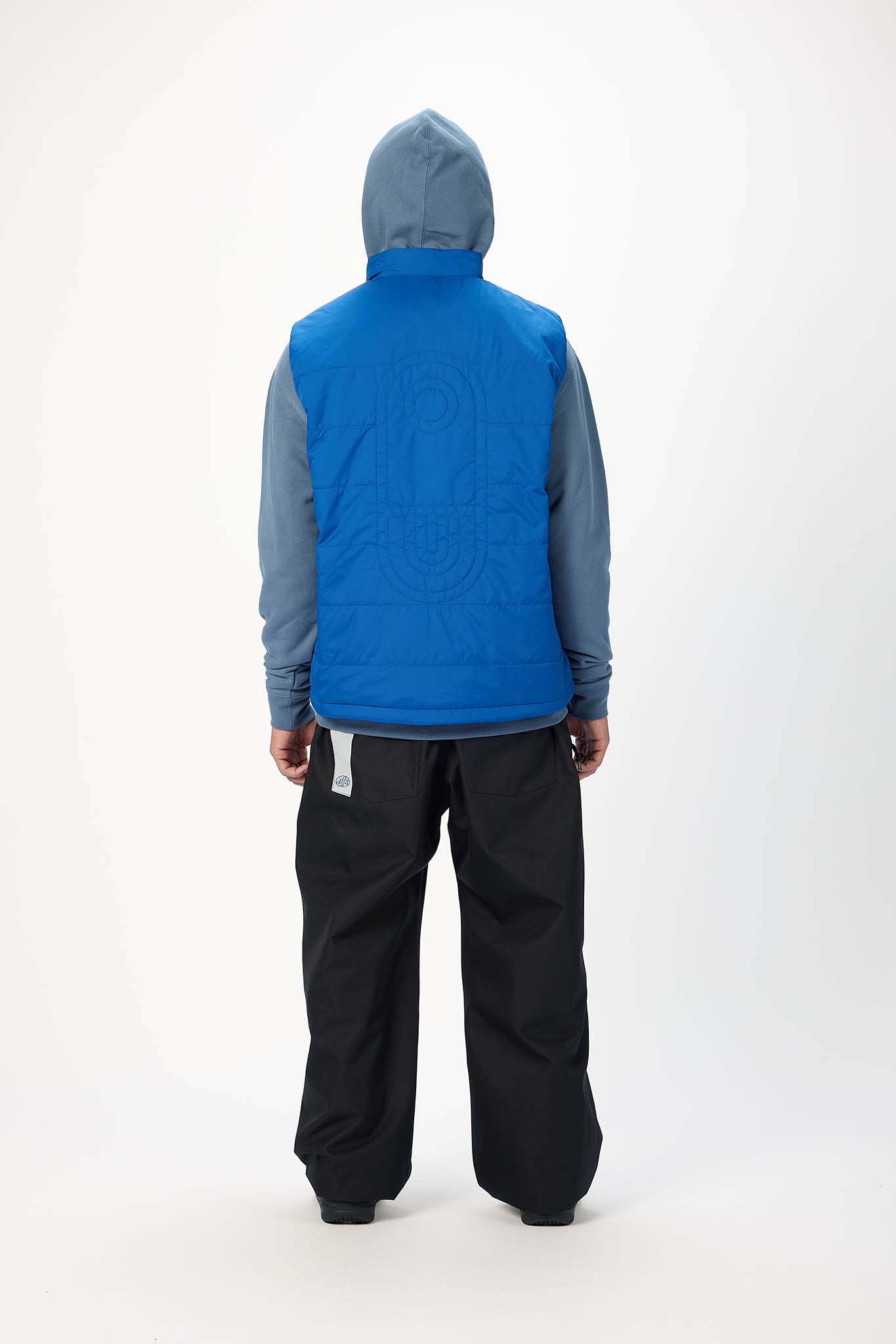 A person shown from the back wearing a bright blue quilted hooded puffer vest over a slate blue long-sleeve top and black snow pants, photographed against a clean white studio background.