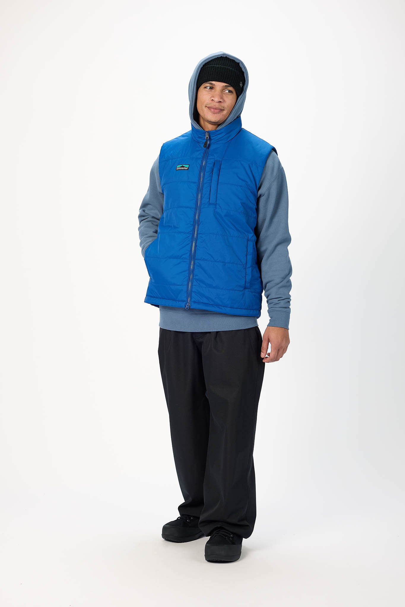 A man wearing a bright blue zip-up puffer vest over a slate blue hoodie, black beanie, and black pants, standing against a plain white studio background.