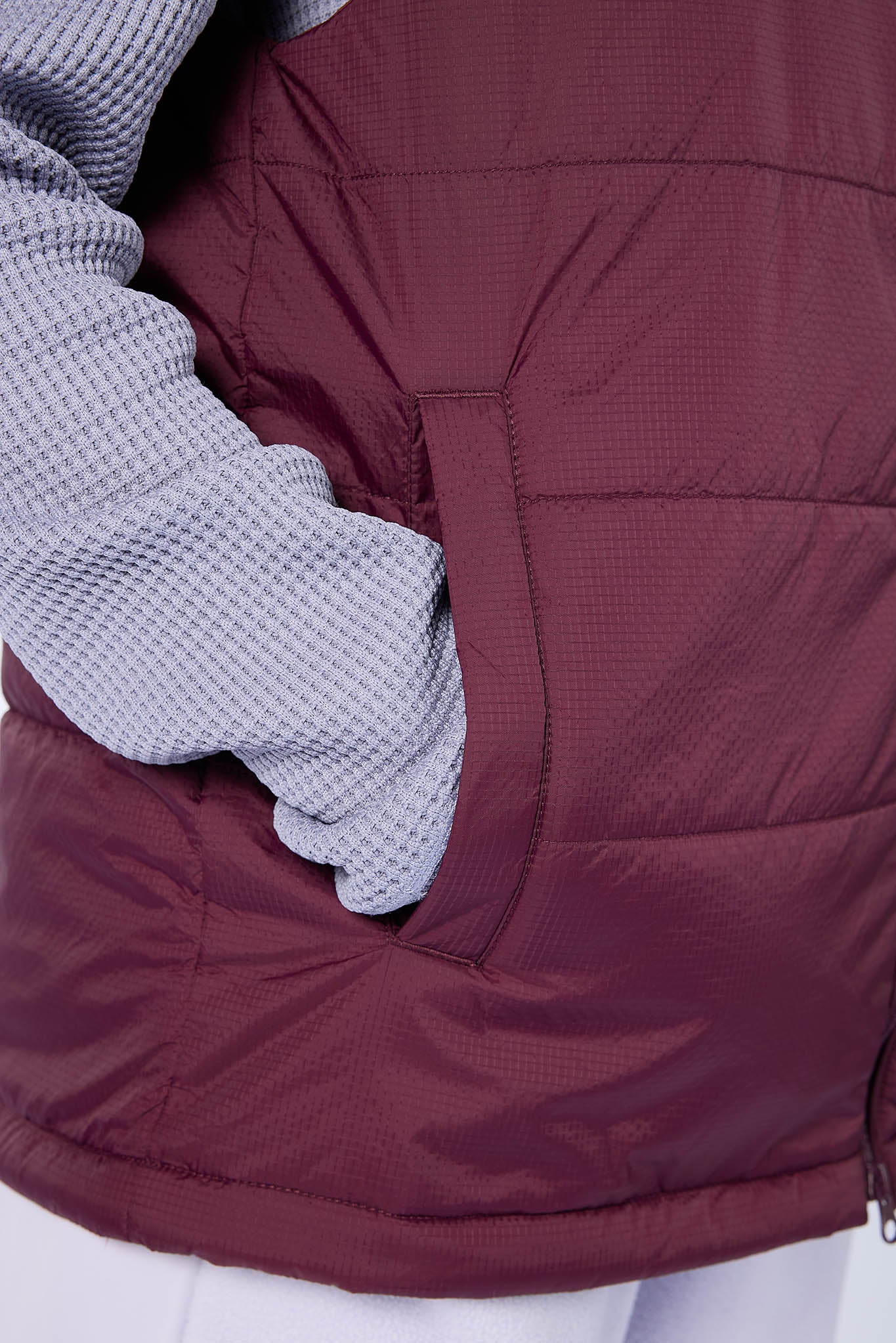 Close-up of a burgundy quilted puffer vest worn over a light gray waffle-knit sweater, showing the side pocket with a hand inside.