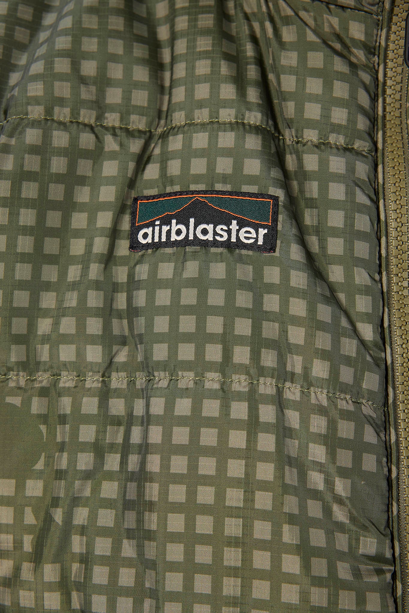 Close-up of a puffer jacket interior showing a green and beige checkered quilted lining with a black Airblaster brand patch.