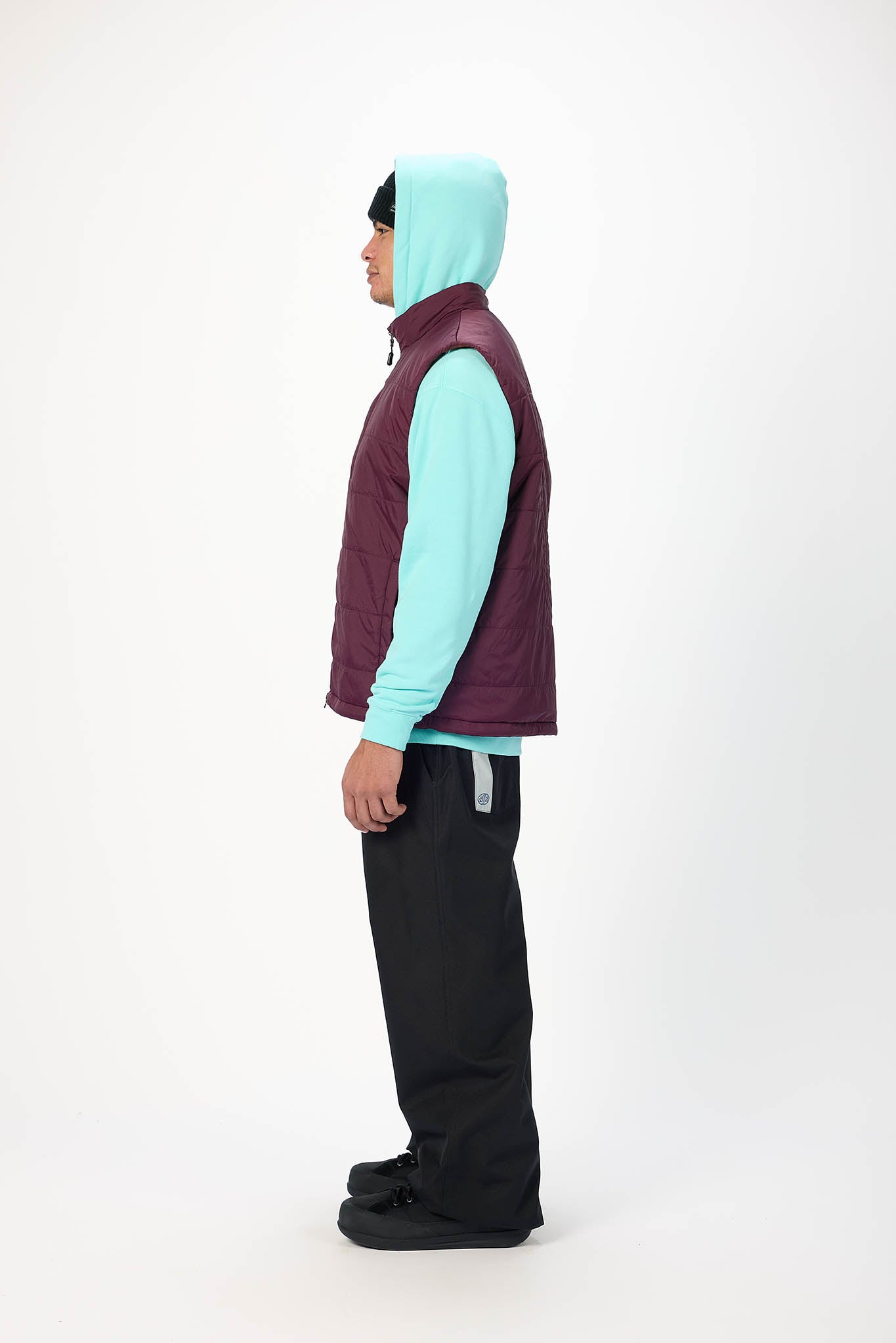 A person shown in profile wearing a dark plum quilted puffer vest over a light aqua hooded sweatshirt, black pants, and black shoes, photographed against a plain white studio background.