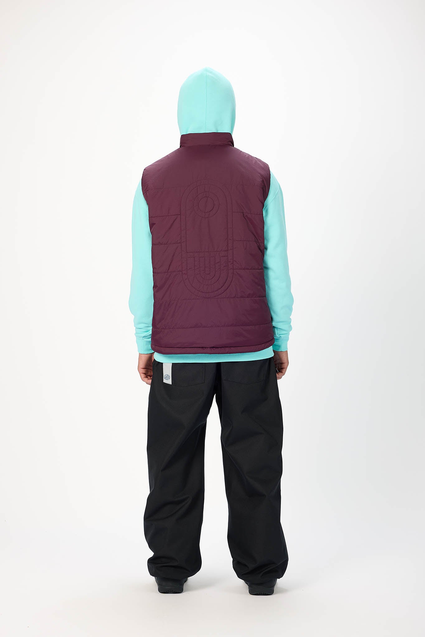 A model shown from the back wearing a maroon quilted puffer vest over a light aqua hooded sweatshirt and black pants, against a plain white studio background.