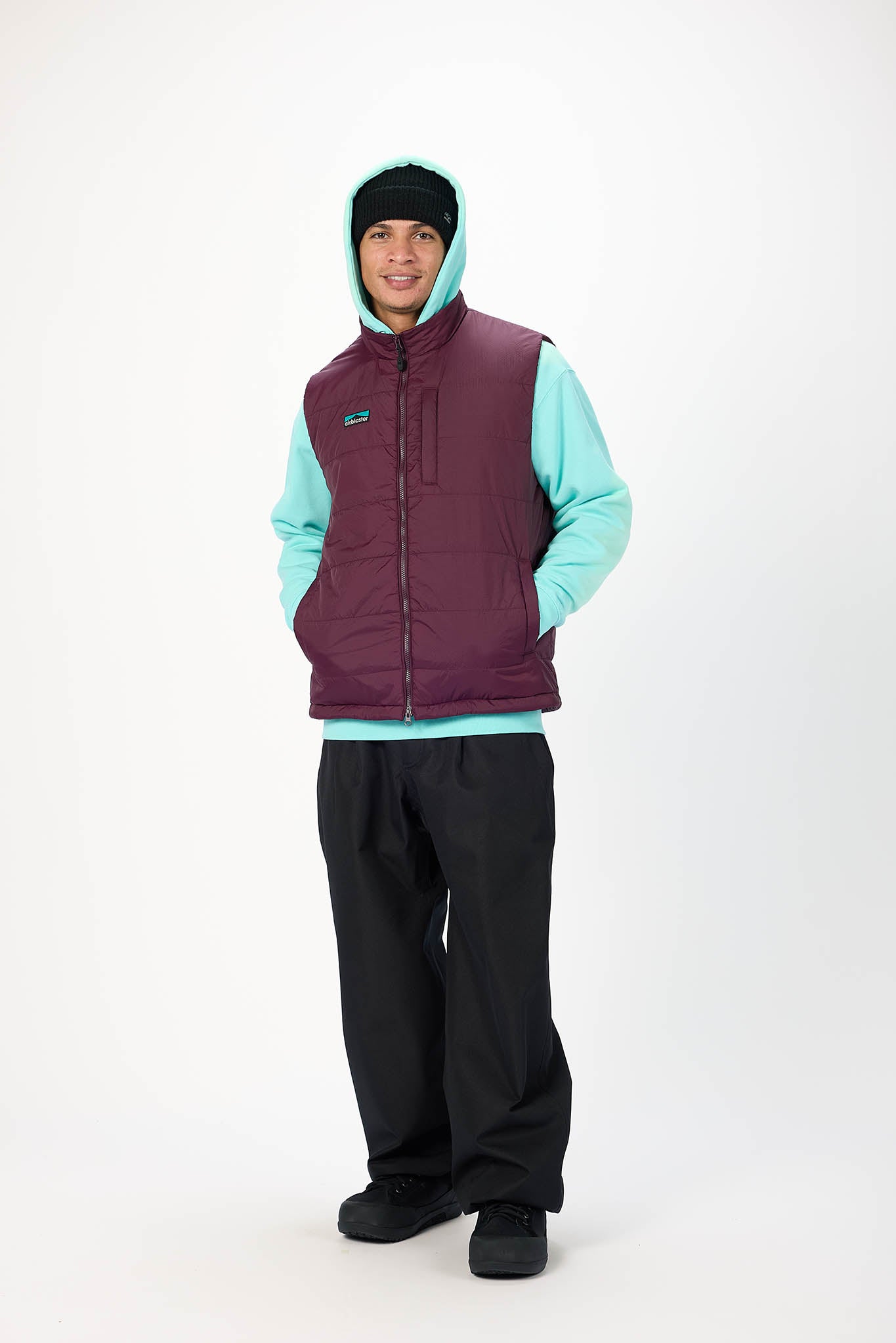A model wearing a burgundy puffer vest over a light teal hoodie, black pants, and a black beanie, standing against a plain white studio background.