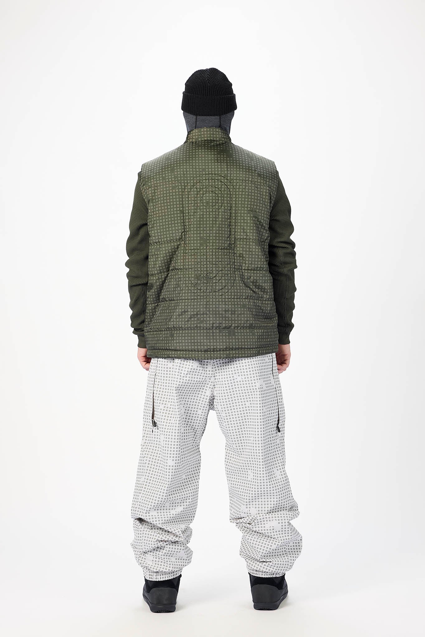 A model shown from the back wearing a dark olive green quilted jacket, light gray and white checkered snow pants, a black beanie, and black boots, set against a plain white studio background.