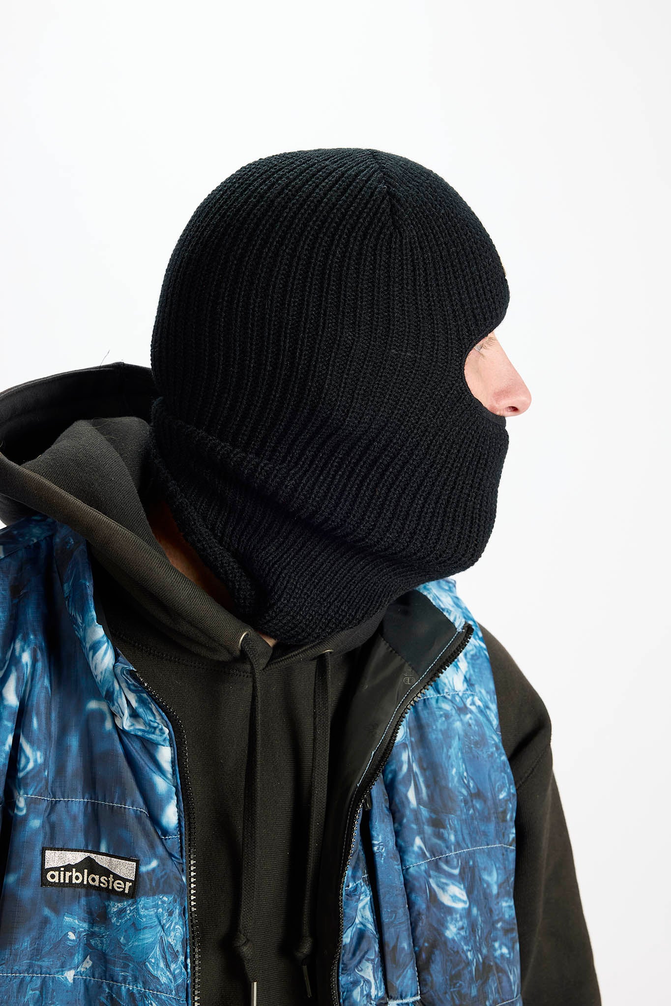 A person wearing a black ribbed knit balaclava mask over the head and neck, paired with a black and blue patterned jacket. The person is shown in profile against a plain white background.