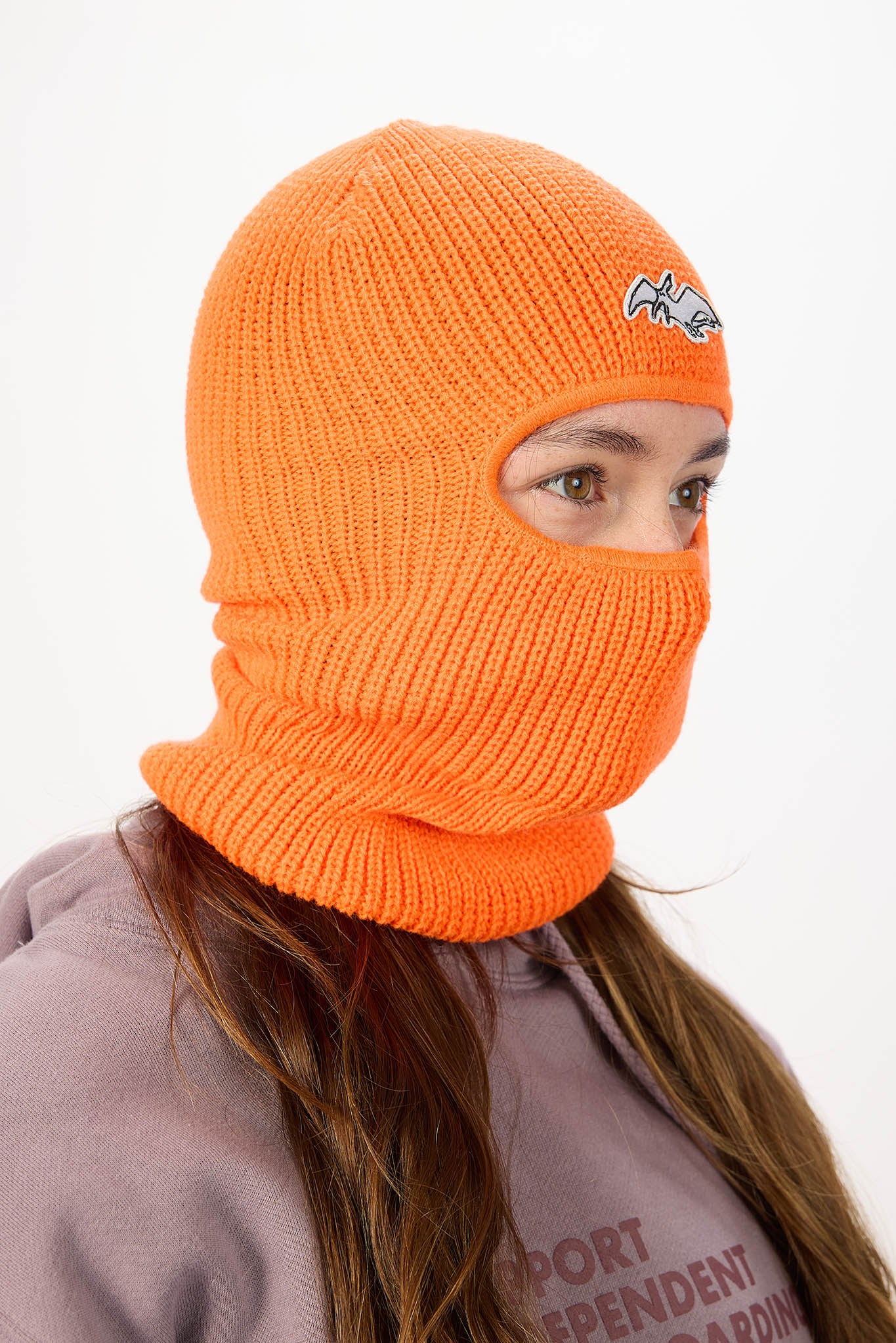A bright orange knit balaclava hood with a small white embroidered logo on the forehead, worn by a model against a plain white background.