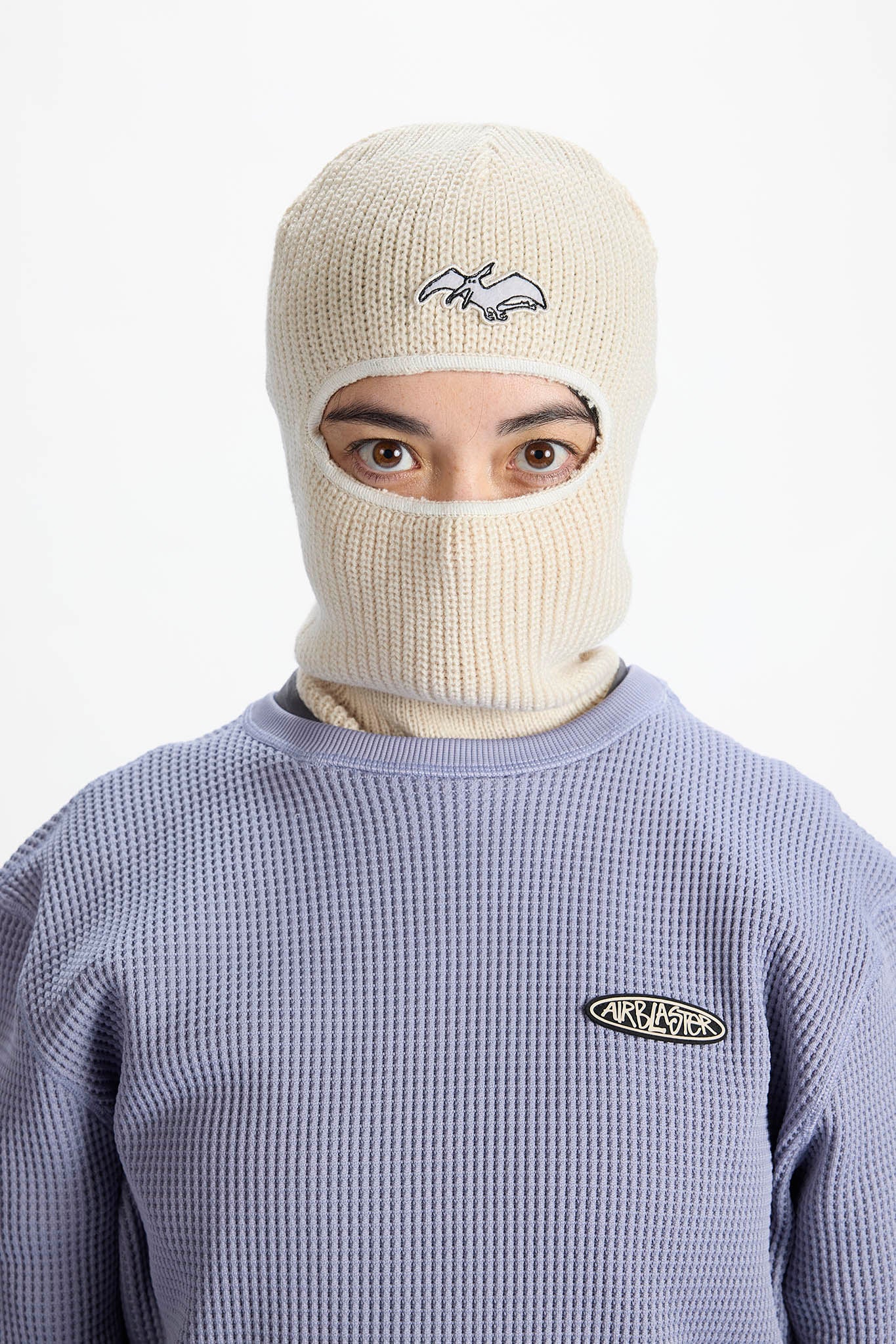 A model wearing a cream-colored ribbed knit balaclava mask with a small embroidered logo, paired with a lavender waffle-knit sweatshirt. The background is a clean, solid white studio backdrop.