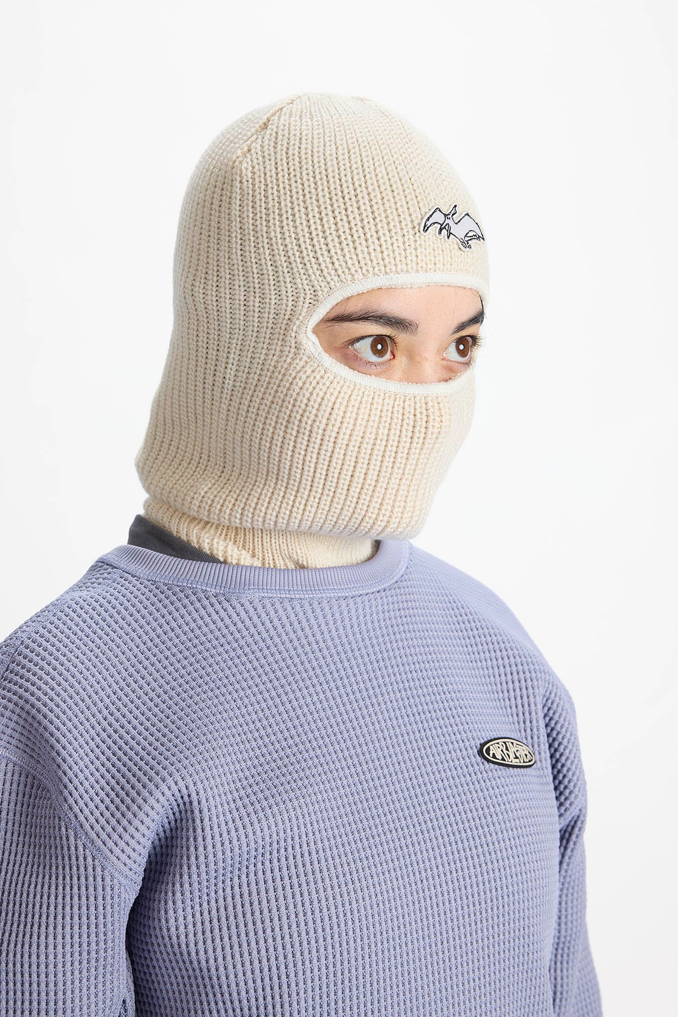 A person wearing a cream-colored ribbed knit balaclava mask with a small embroidered logo, paired with a lavender waffle-knit sweatshirt. The background is a clean, solid white studio backdrop.