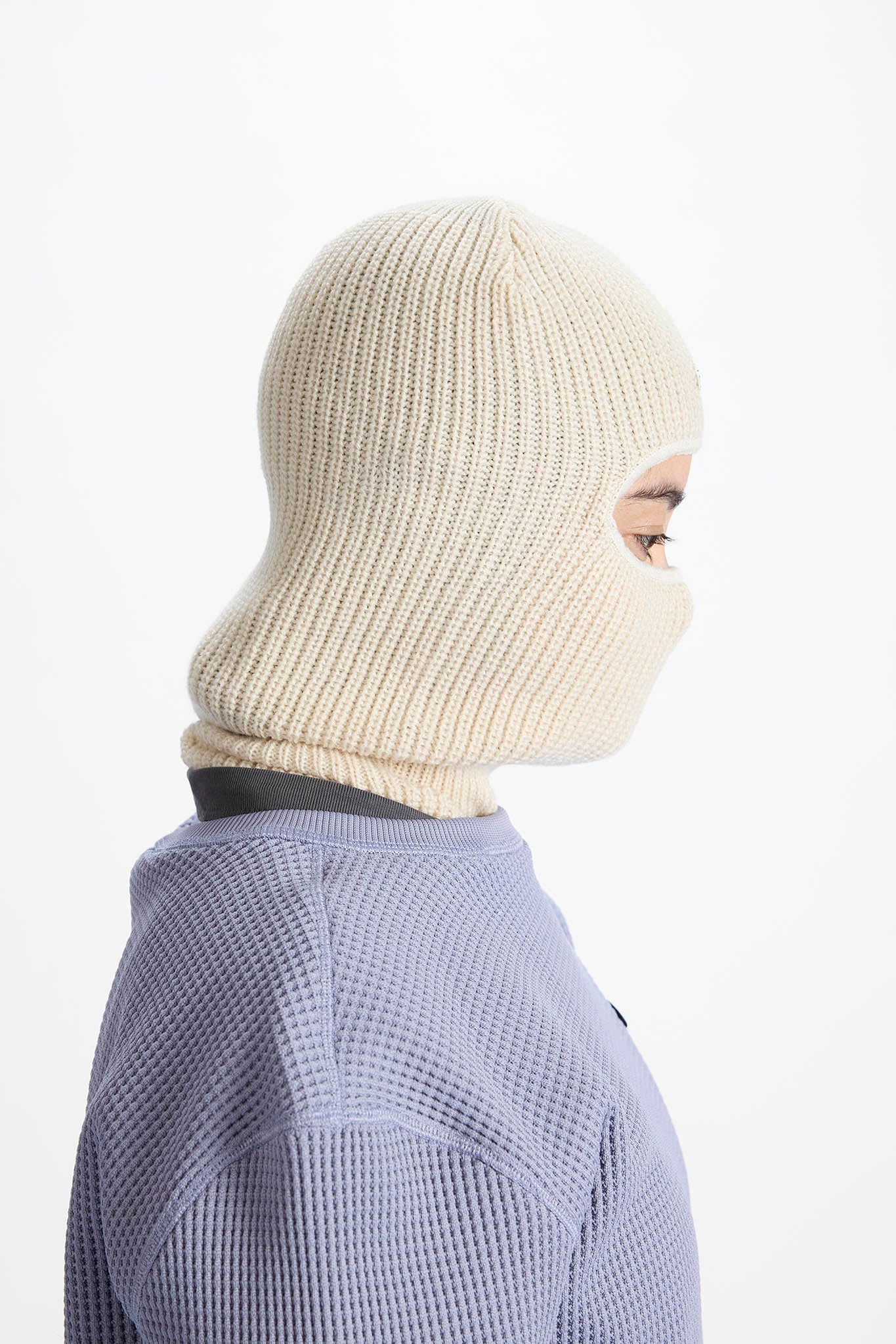 A side profile view of a person wearing a cream-colored ribbed knit balaclava mask and a lavender waffle-knit top against a plain white background.