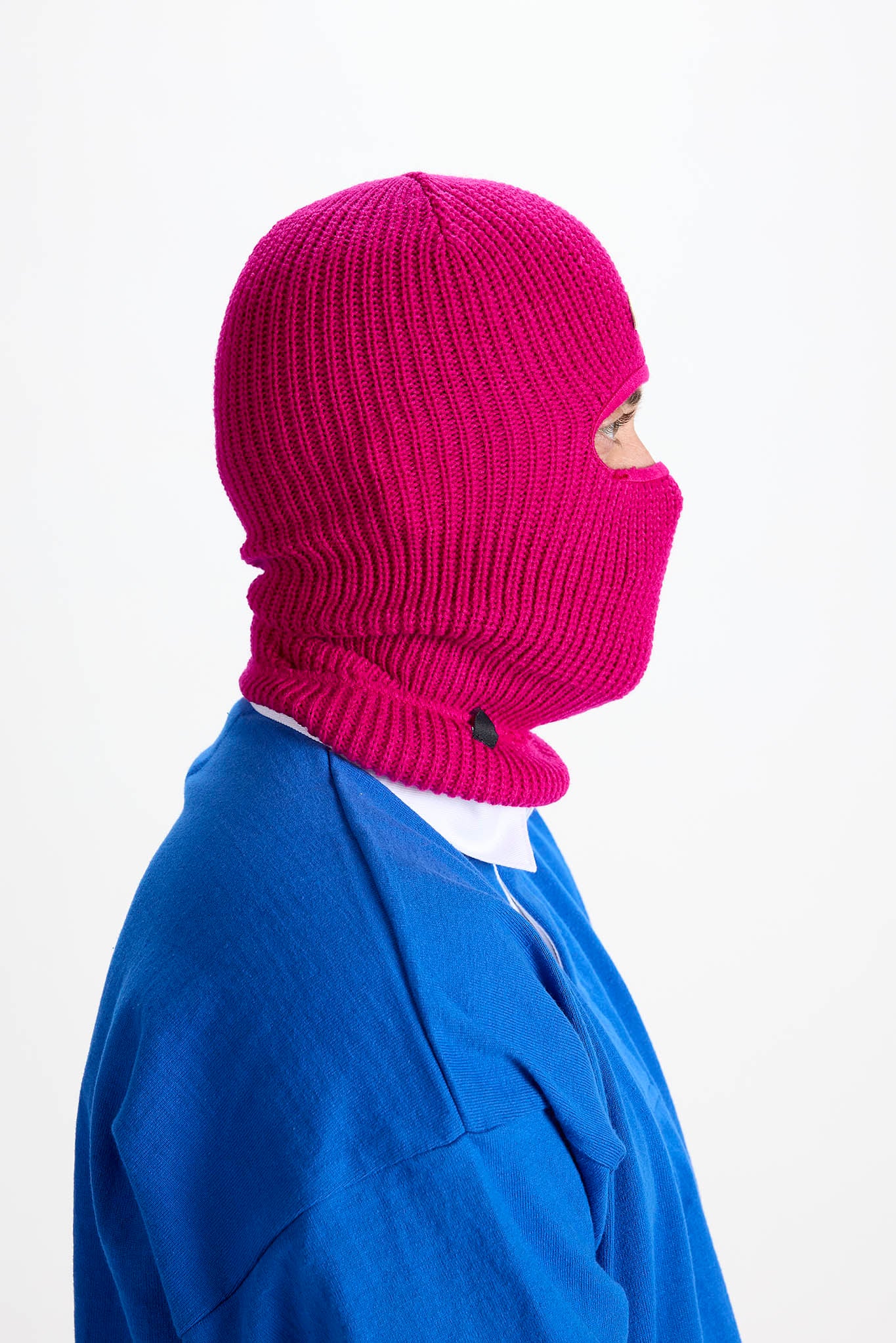 A person in profile wearing a bright magenta ribbed knit balaclava mask over a blue top, photographed against a plain white background.