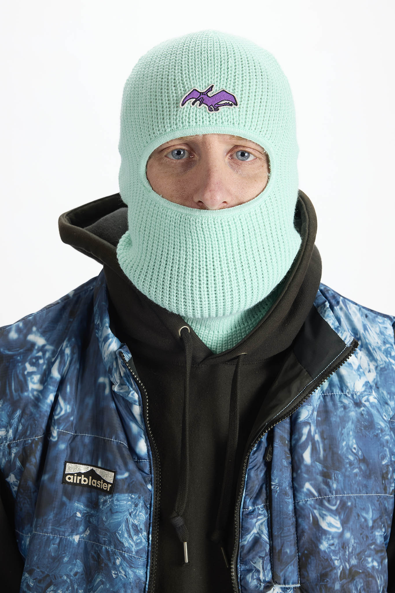 A person wearing a bright teal knit balaclava mask with a small embroidered logo on the forehead, paired with a blue polo shirt against a plain white background.