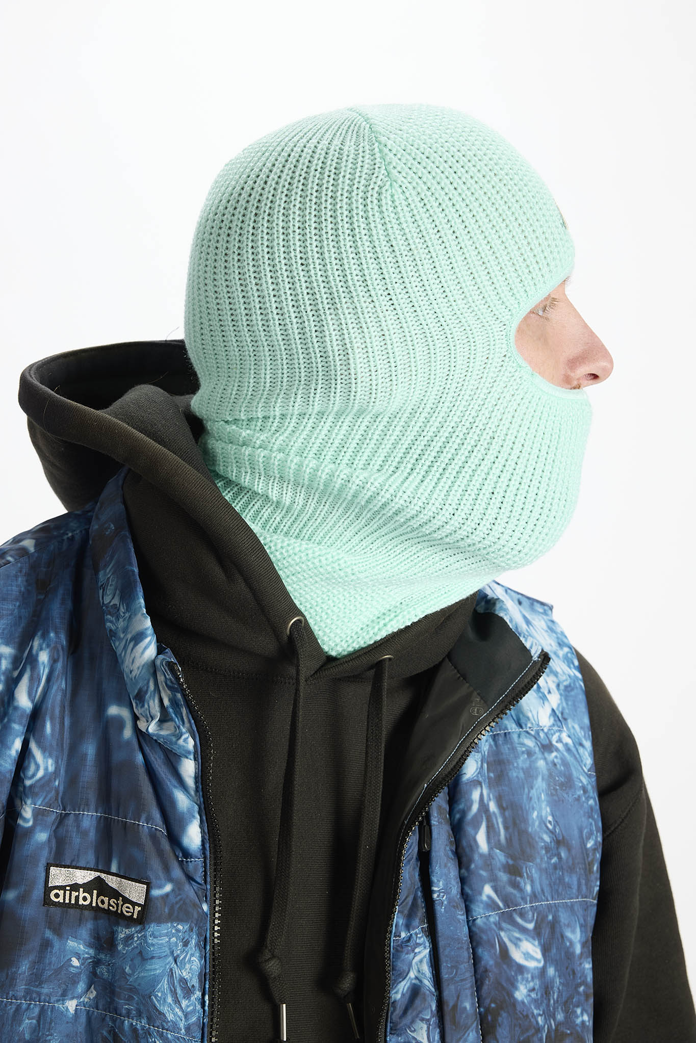A person wearing a mint green ribbed knit balaclava mask, a black hoodie, and a blue patterned Airblaster jacket, shown in profile against a plain white background.