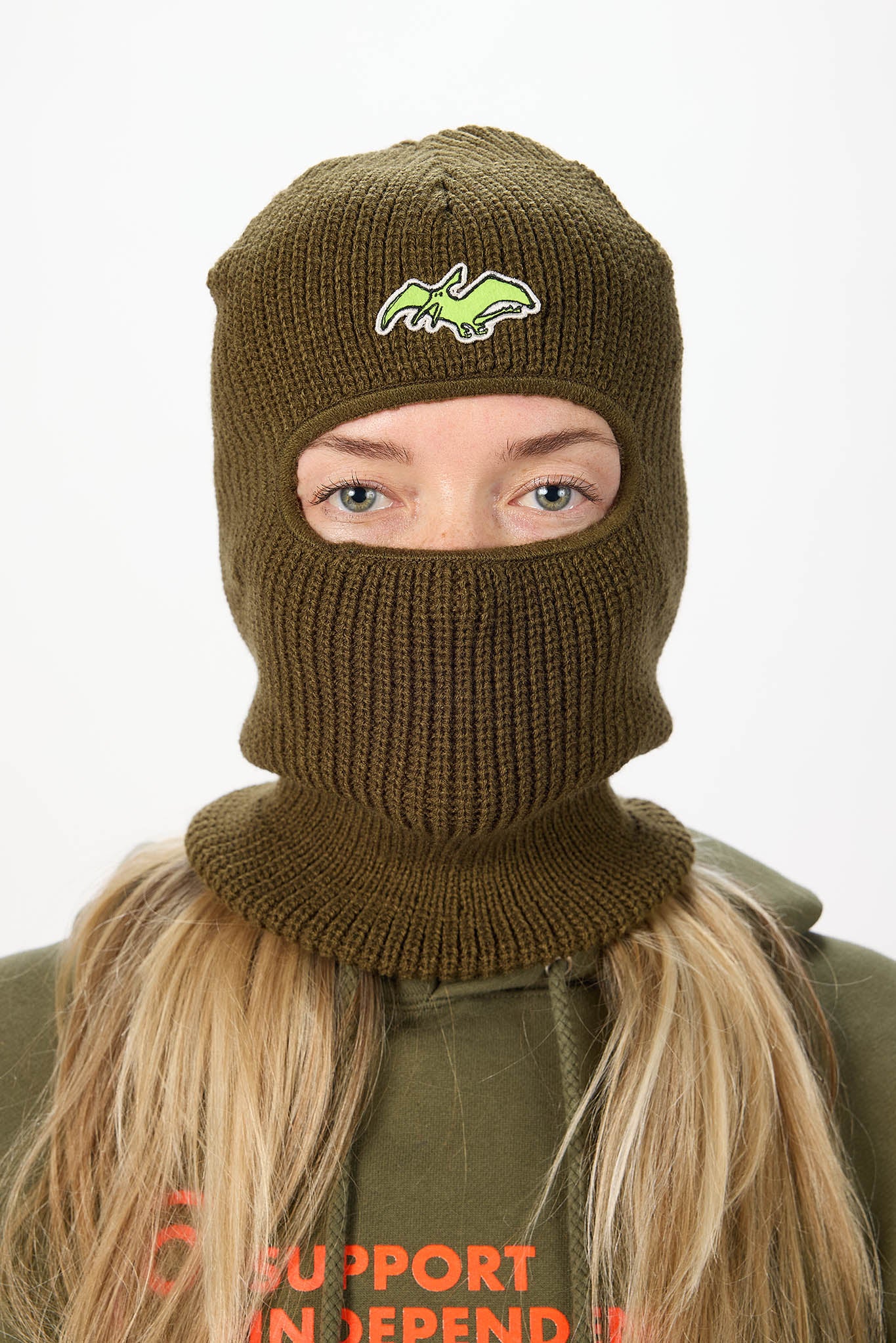 A person wearing an olive green ribbed knit balaclava mask with a small green and white embroidered logo on the forehead, paired with an olive green hoodie against a plain white background.