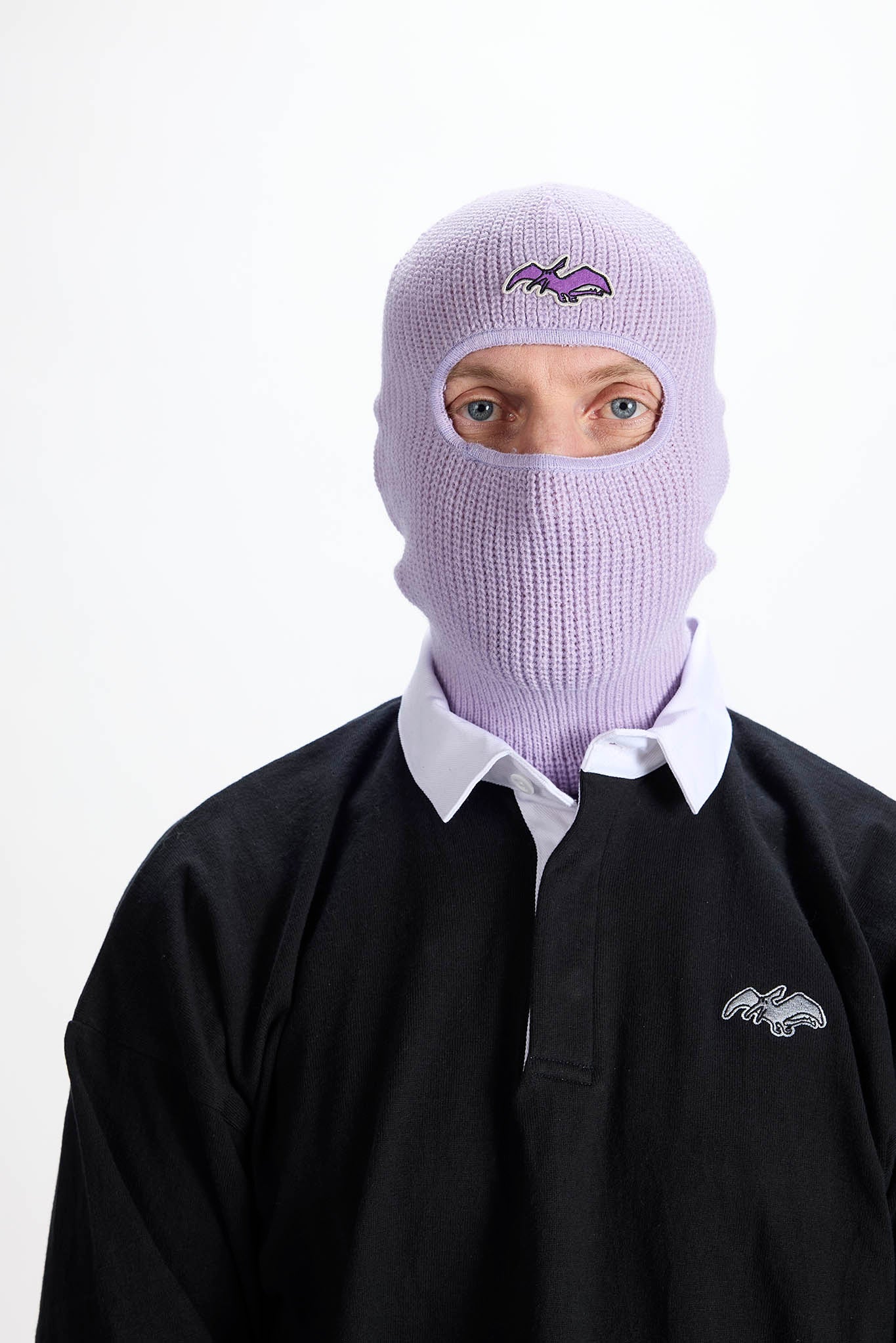 A person wearing a light purple knit balaclava with a small dark bat logo on the forehead, layered over a white collared shirt and a black long-sleeve top with a matching bat logo on the chest, against a plain white background.