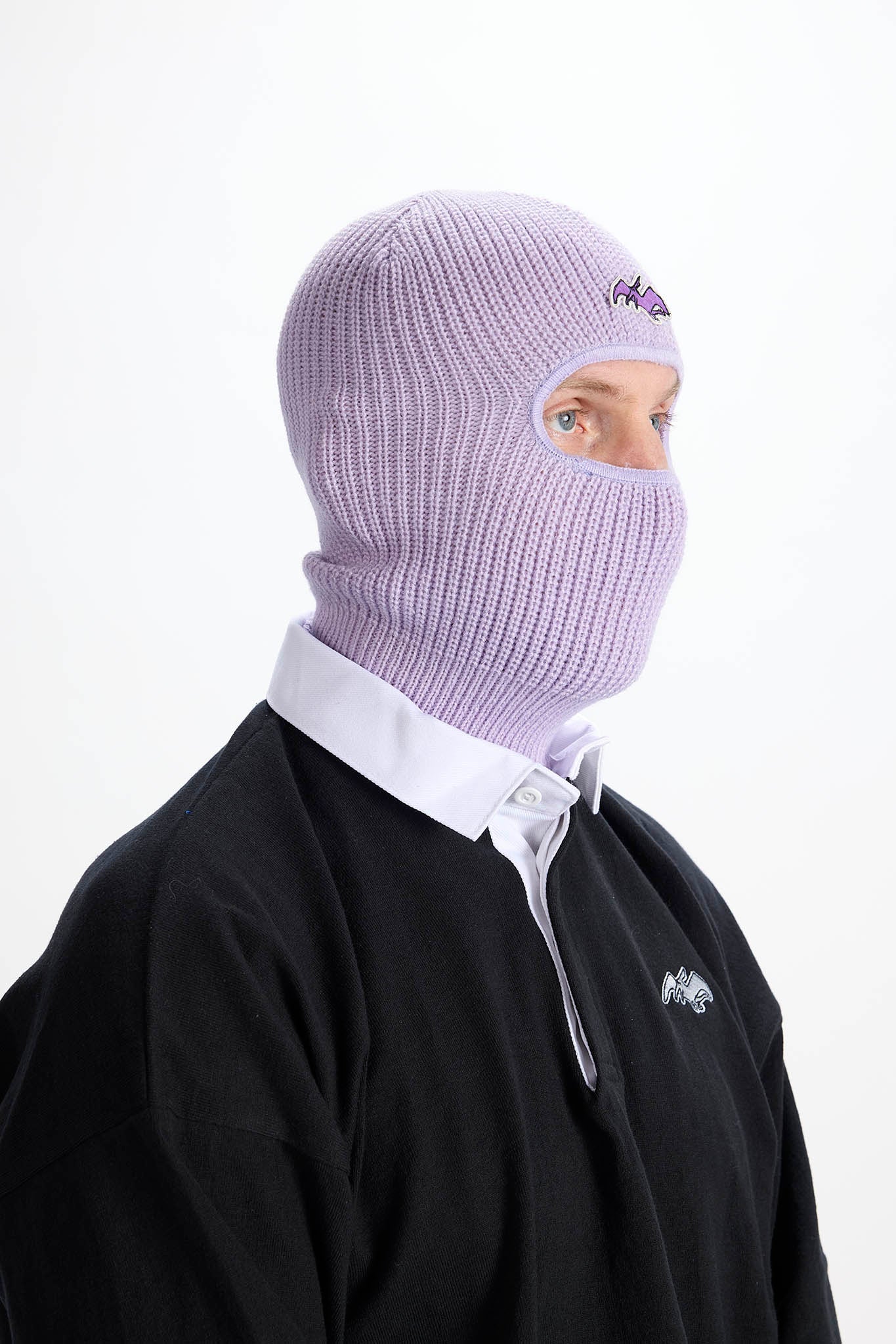 A person wearing a light purple ribbed knit balaclava mask over a black sweatshirt and white collared shirt, shown against a plain white background.
