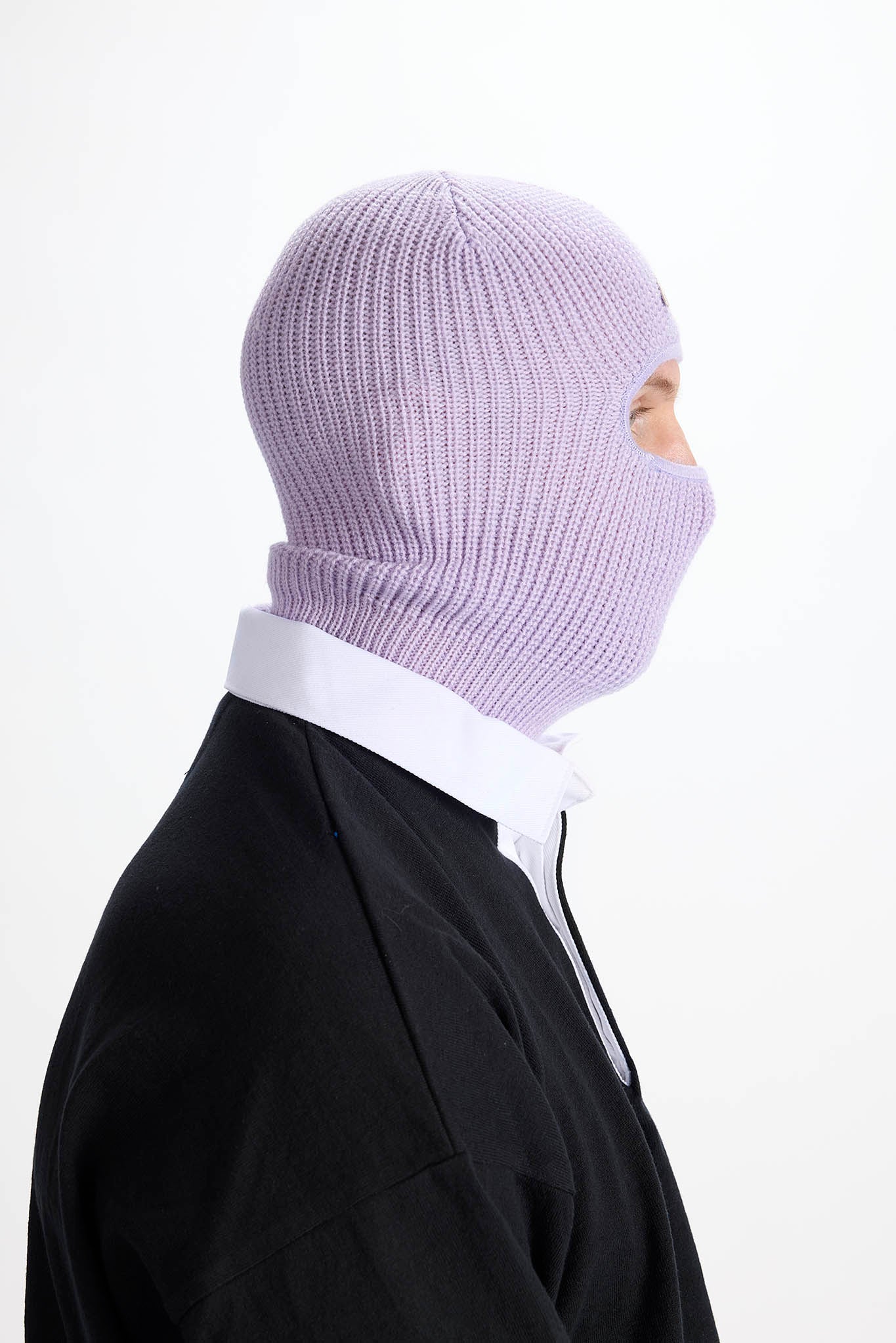 A person shown in side profile wearing a light lavender ribbed knit balaclava mask over a white collared shirt and black jacket, against a plain white background.
