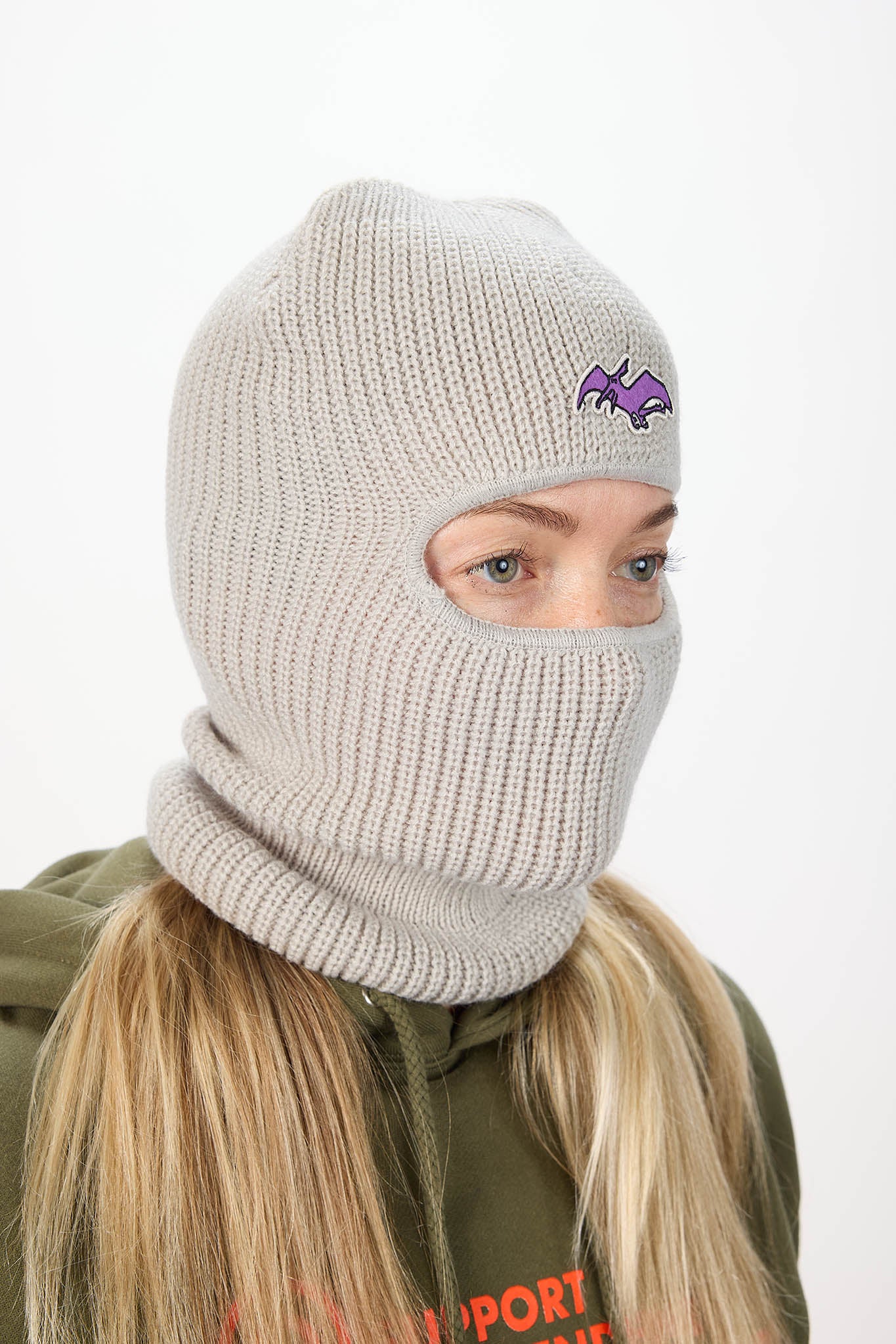 A person wearing a light beige ribbed knit balaclava mask with a small purple bat logo on the forehead, paired with an olive green hoodie against a plain white background.