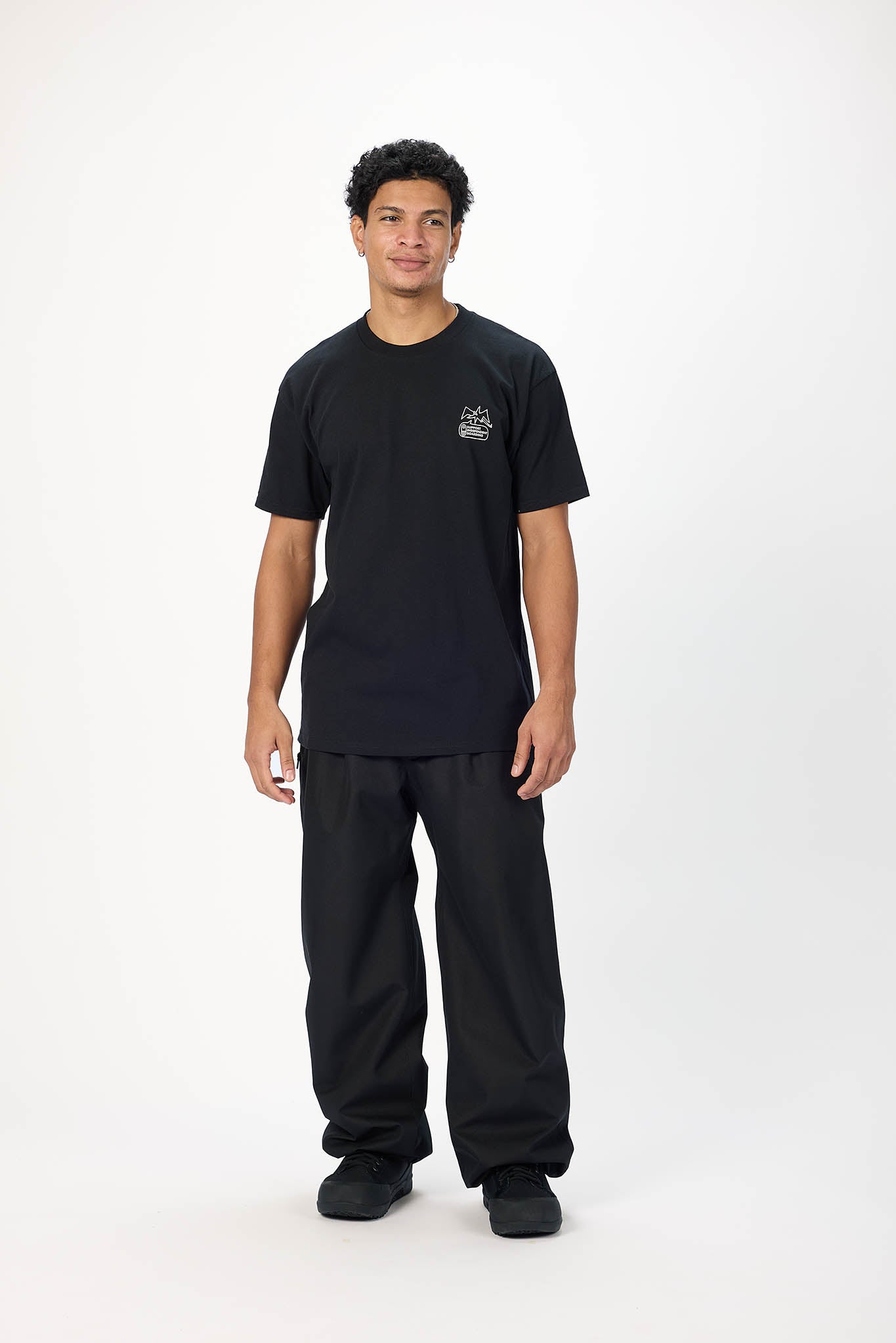 A man modeling a black short-sleeve t-shirt with a small white graphic on the left chest, paired with black pants and black shoes, standing against a plain white studio background.