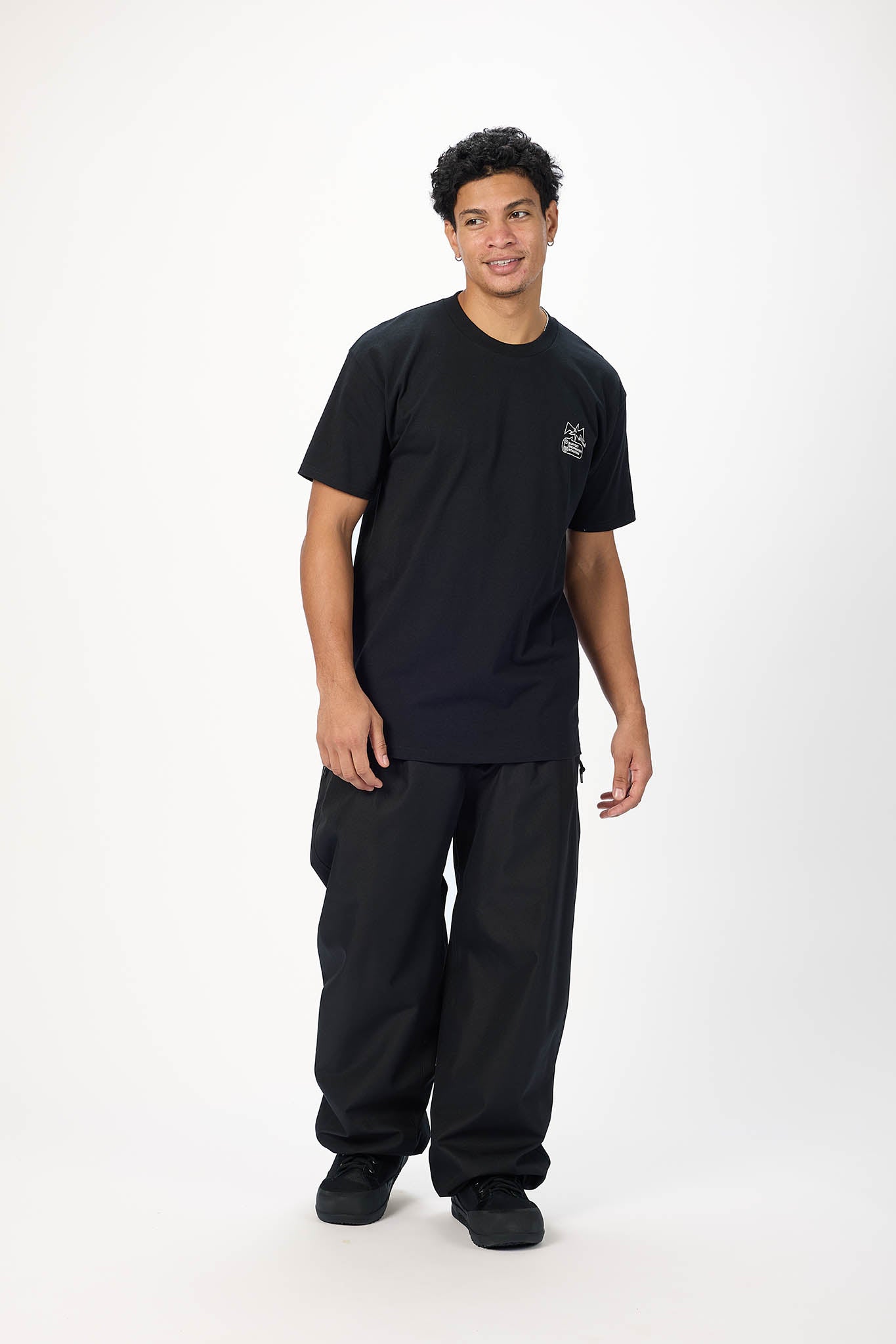 A man modeling a black short-sleeve crewneck t-shirt with a small white graphic on the left chest, paired with black cargo pants, standing against a plain white studio background.