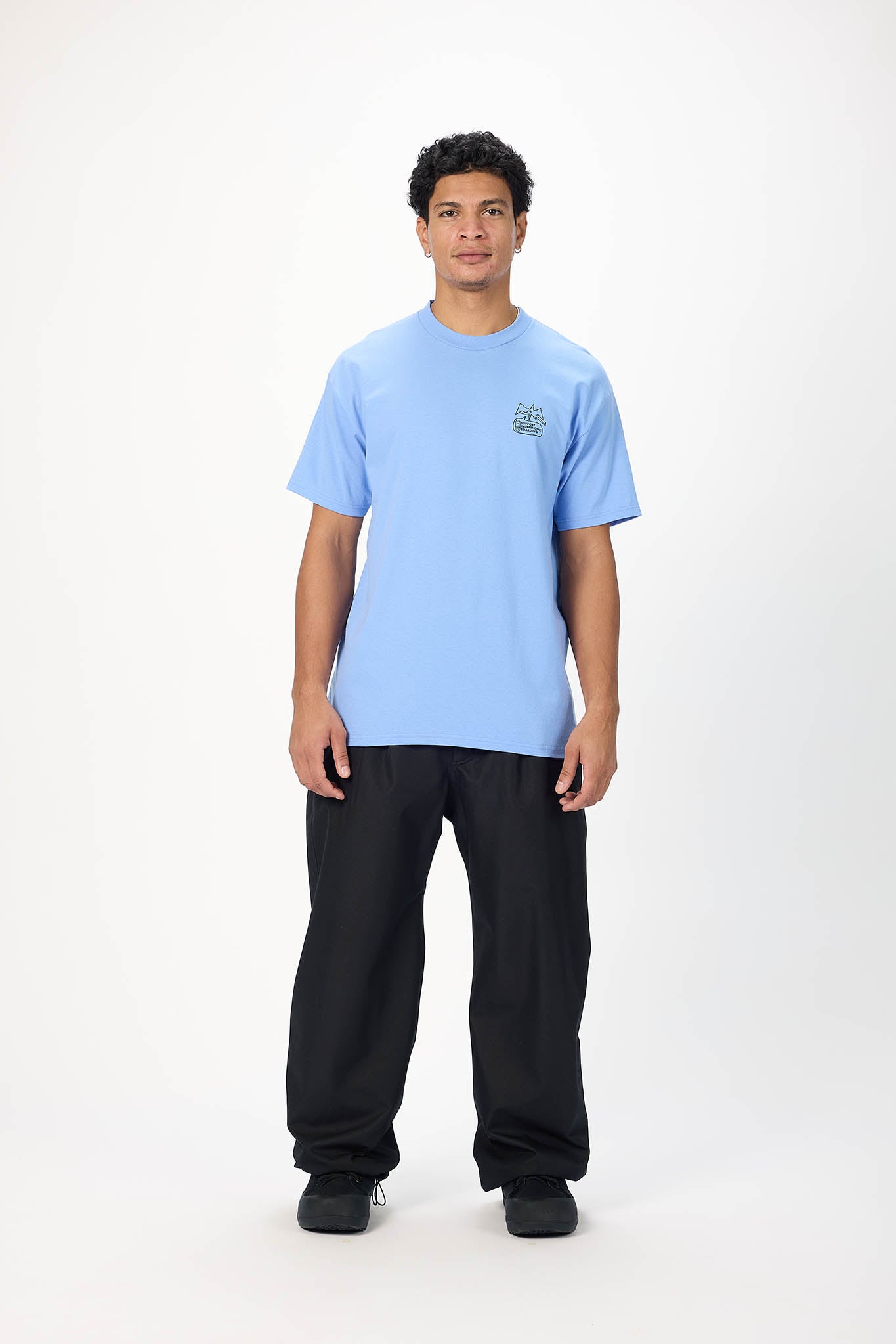 A man modeling a light blue crewneck t-shirt with a small green graphic on the left chest, paired with black pants, standing against a plain white studio background.