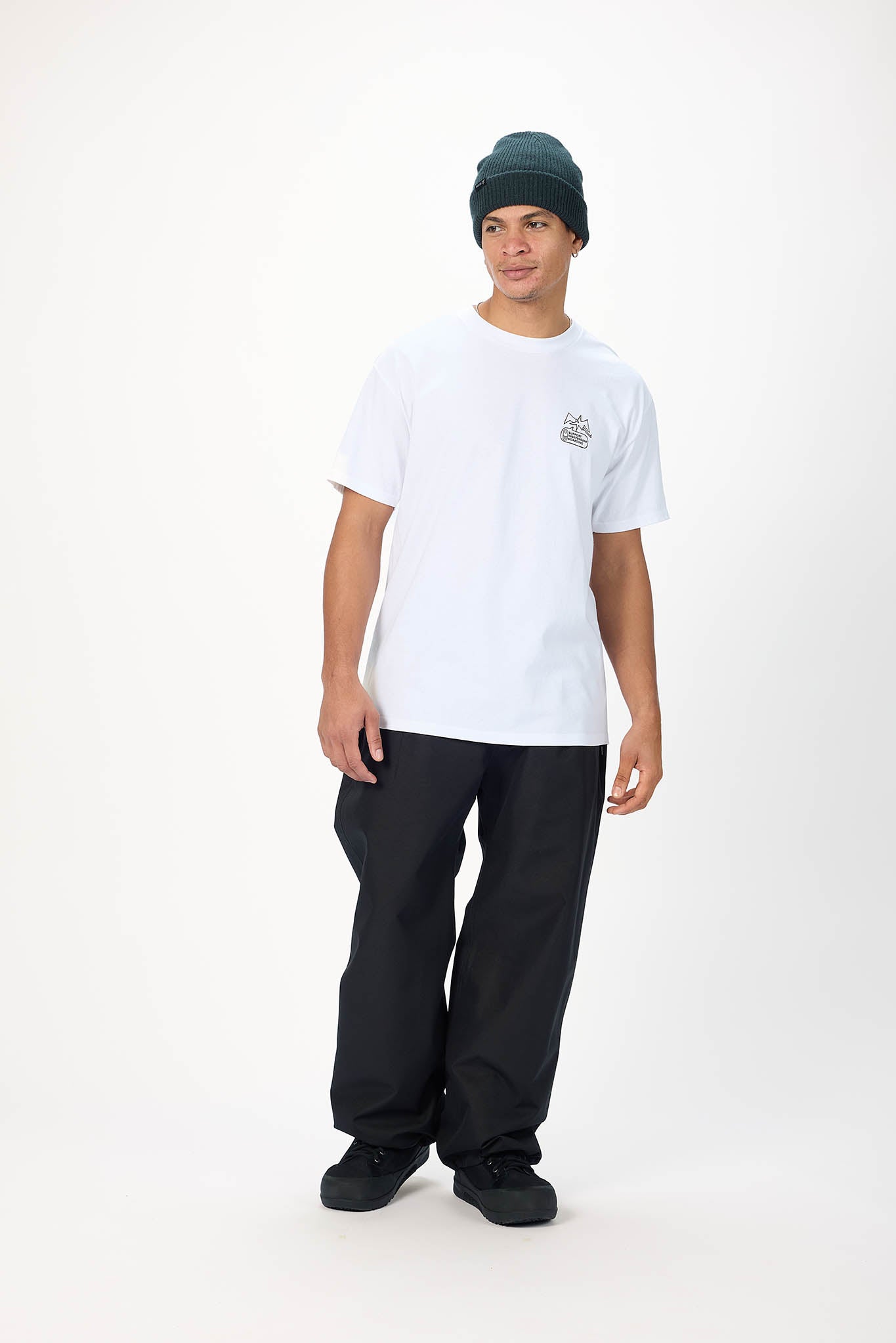 A man modeling a white short-sleeve t-shirt with a small graphic on the left chest, paired with black pants and a dark green beanie, standing against a plain white studio background.