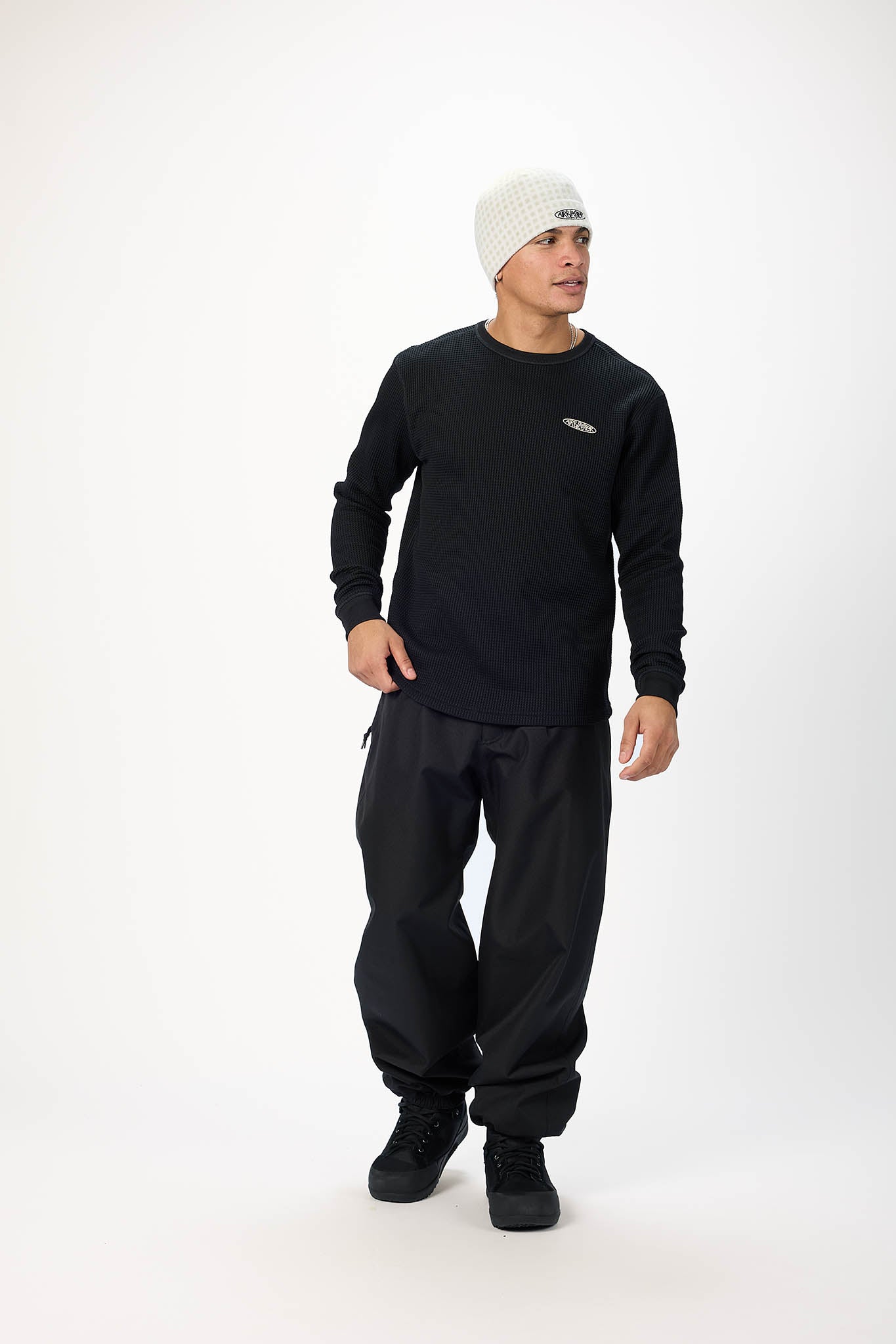 A man wearing a black long-sleeve crewneck sweatshirt, black baggy sweatpants, black sneakers, and a white knit beanie, standing against a plain white studio background.