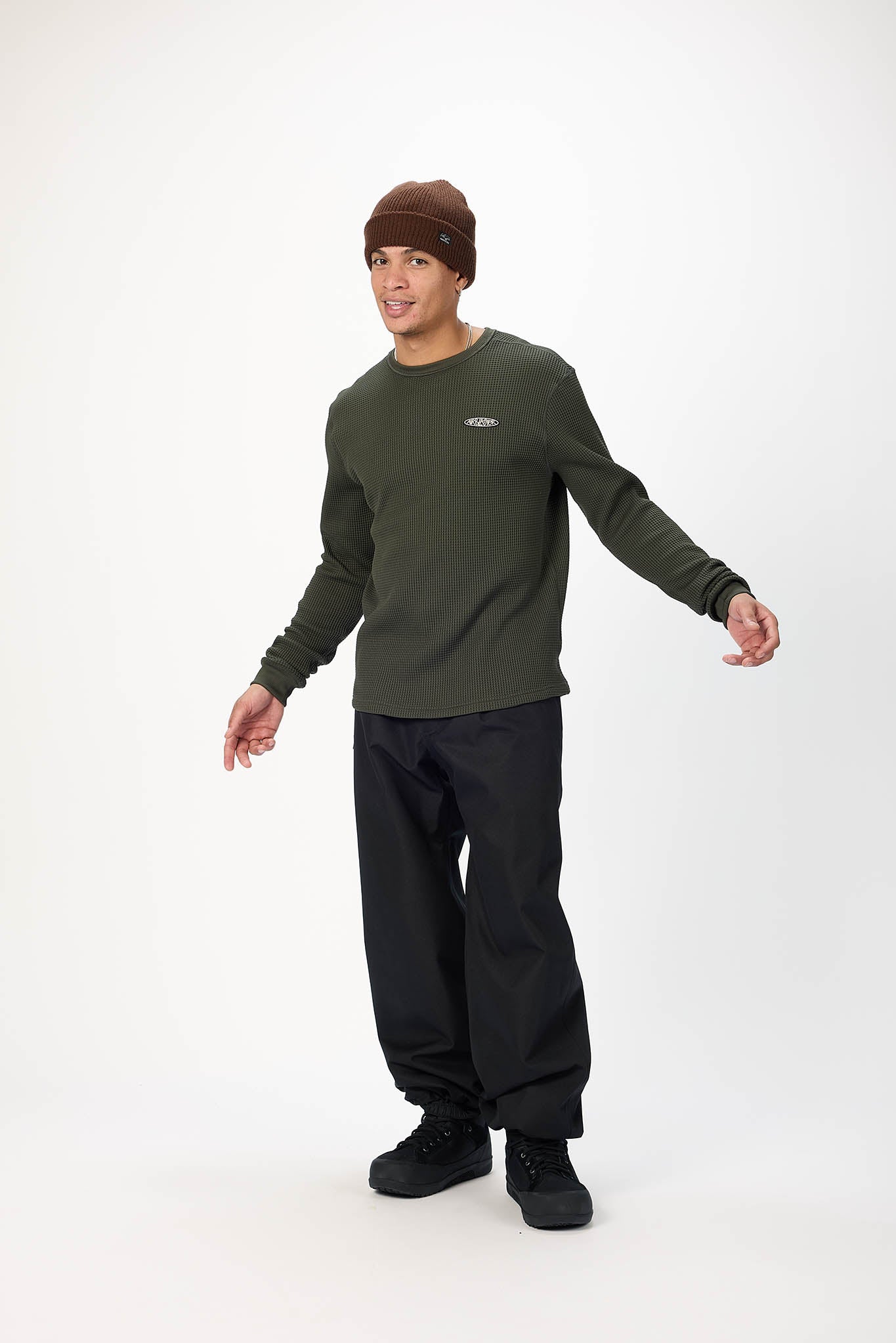 A man modeling a dark olive green long-sleeve crewneck shirt, black pants, black shoes, and a brown beanie against a plain white studio background.