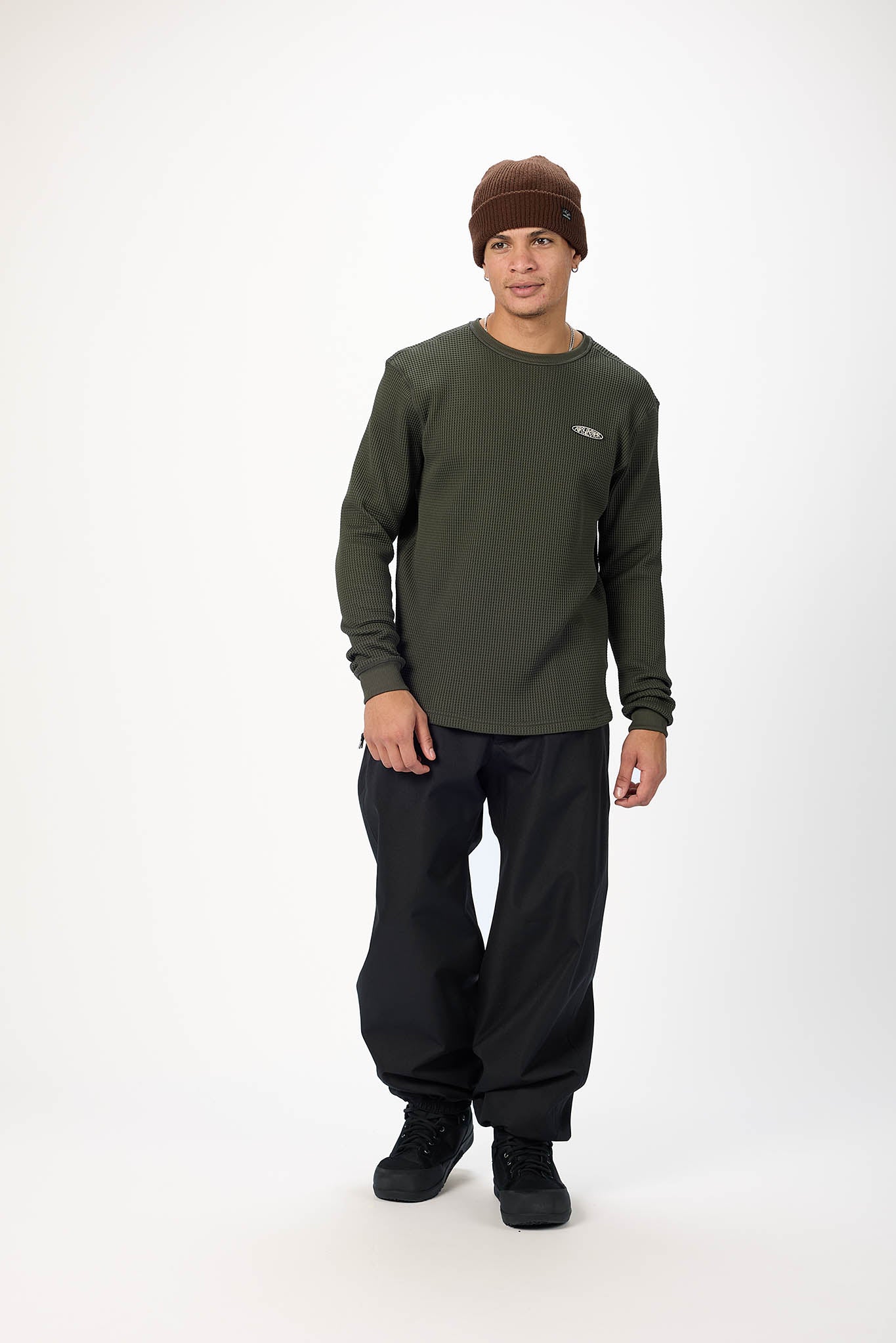 A man modeling a dark olive green long-sleeve crewneck shirt, black pants, black shoes, and a brown knit beanie against a plain white studio background.