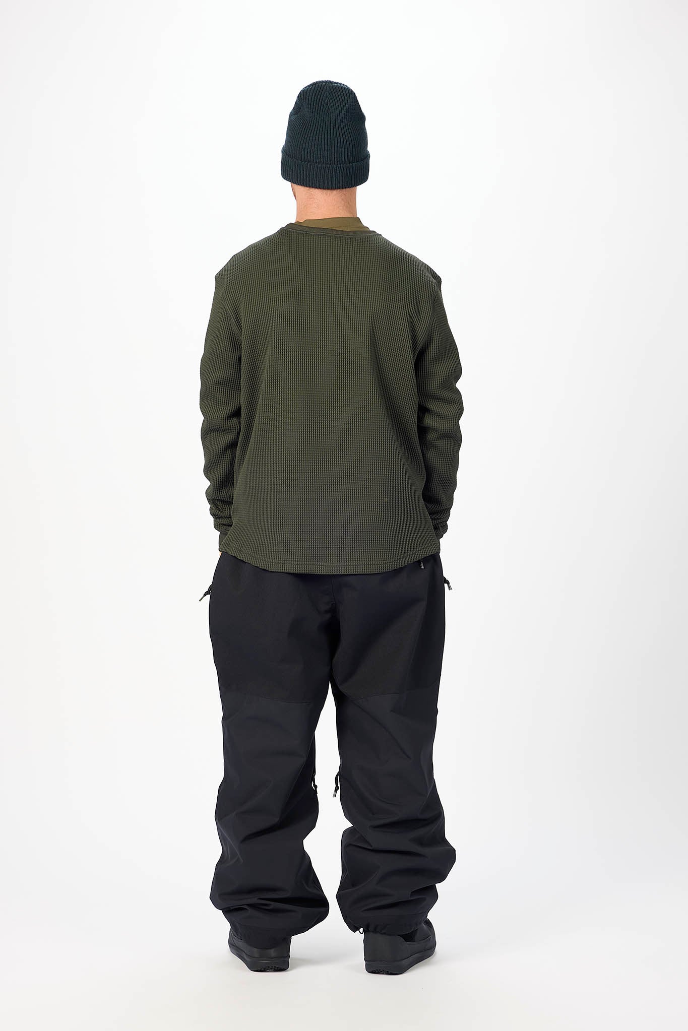 A person shown from the back wearing an olive green long-sleeve shirt, black snow pants, and a black beanie against a plain white studio background.