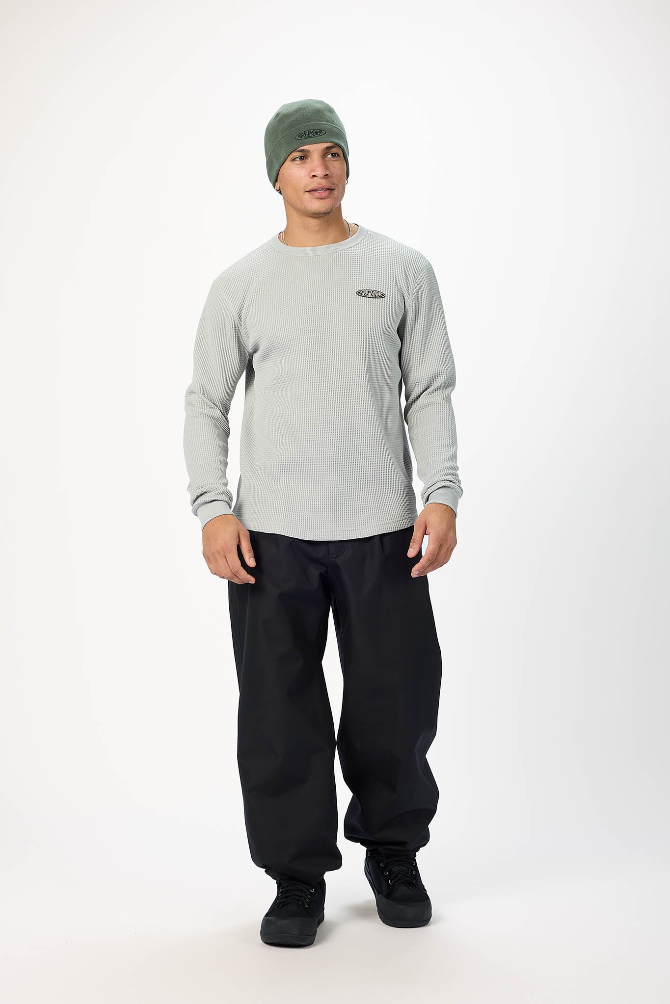 A man modeling a light gray long-sleeve crewneck sweatshirt, black sweatpants, black sneakers, and a green beanie against a plain white studio background.