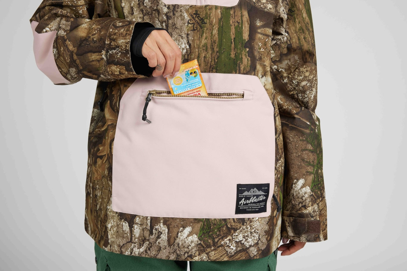 A close-up of a person wearing a camouflage jacket with a large pale pink front pocket. The person is holding a small yellow card into the zippered pocket.