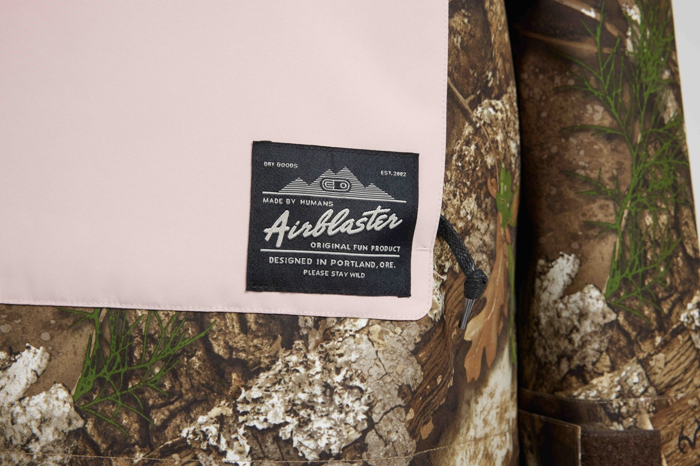 A close-up of a light pink fabric item with a black sewn-in label reading 'Aireblaster' and 'Made by Haulos,' placed over a camouflage-patterned surface with small green plant details.
