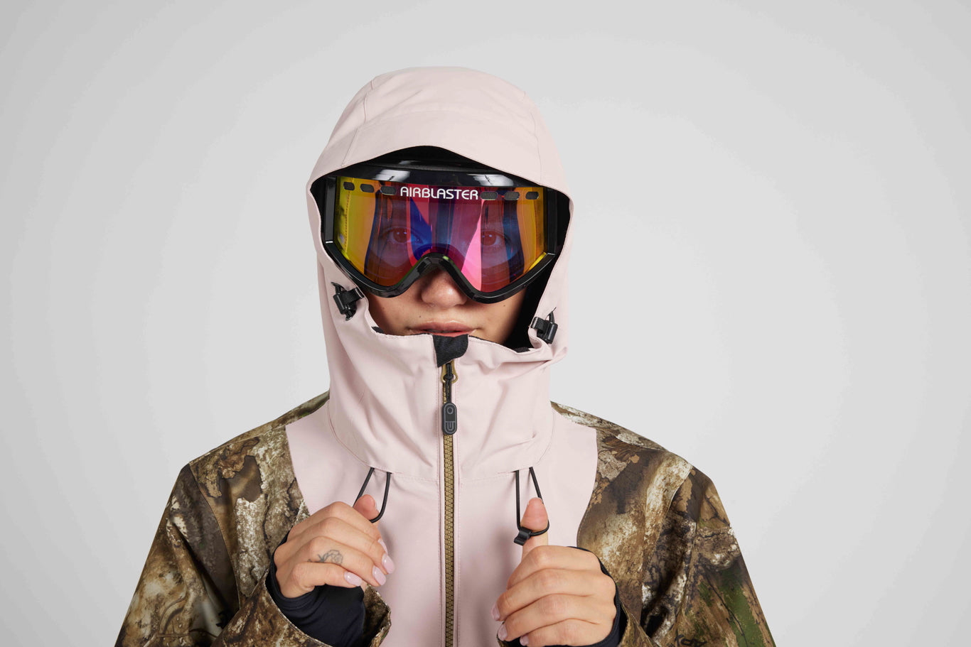A person wearing a light pink ski hood over a camouflage jacket, with black ski goggles featuring a colorful reflective lens. The brand name 'AirBlaster' is visible on the top of the goggles.