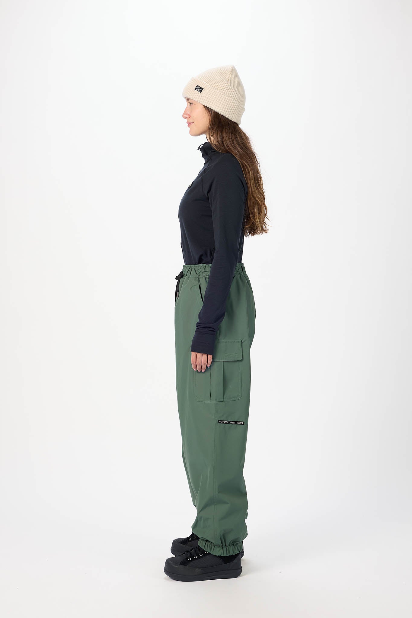 Person wearing a black top, green pants, and a beige beanie on a white background