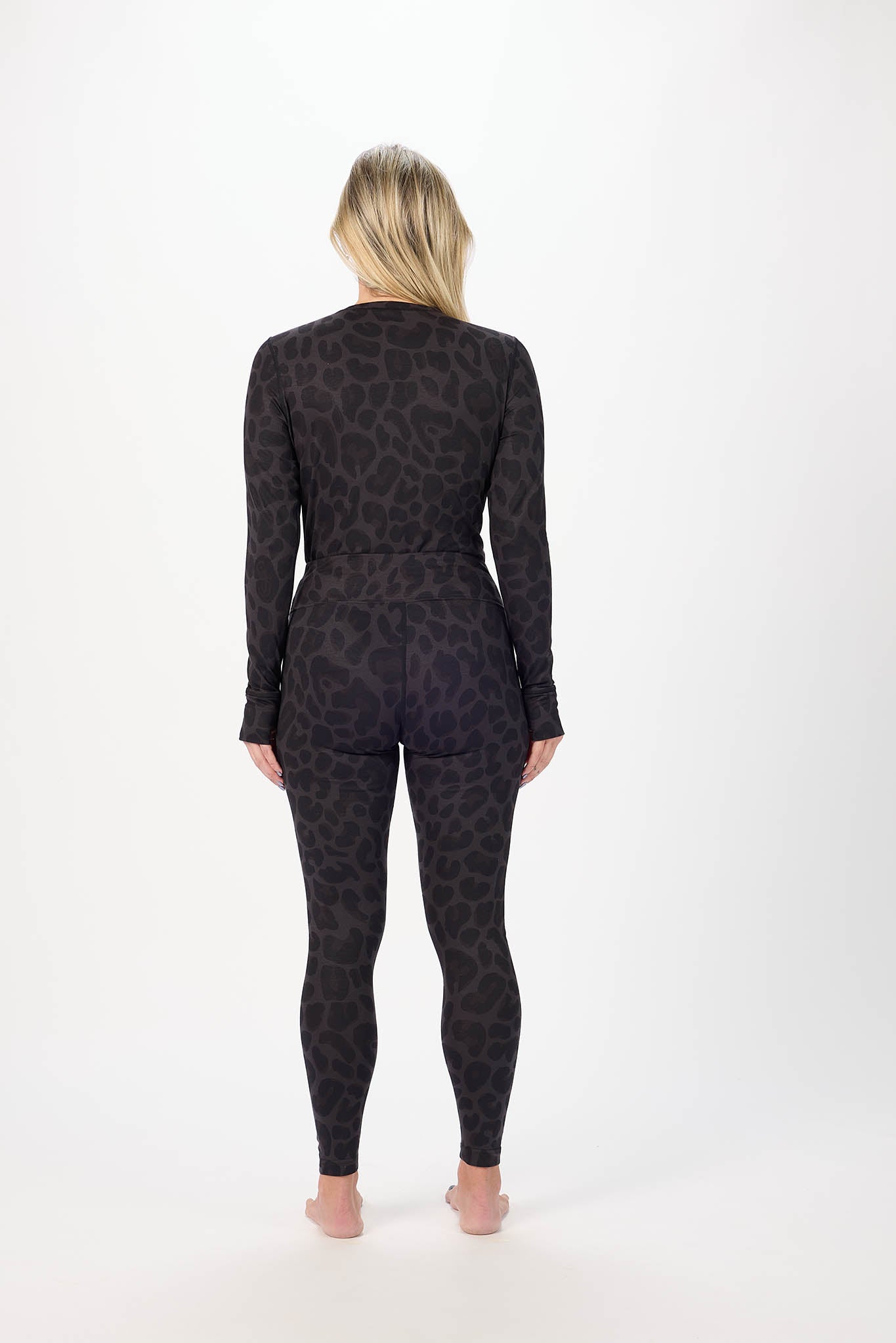 A woman viewed from behind wearing a matching long-sleeve top and leggings set in a dark charcoal gray and black leopard print, standing against a plain white studio background.