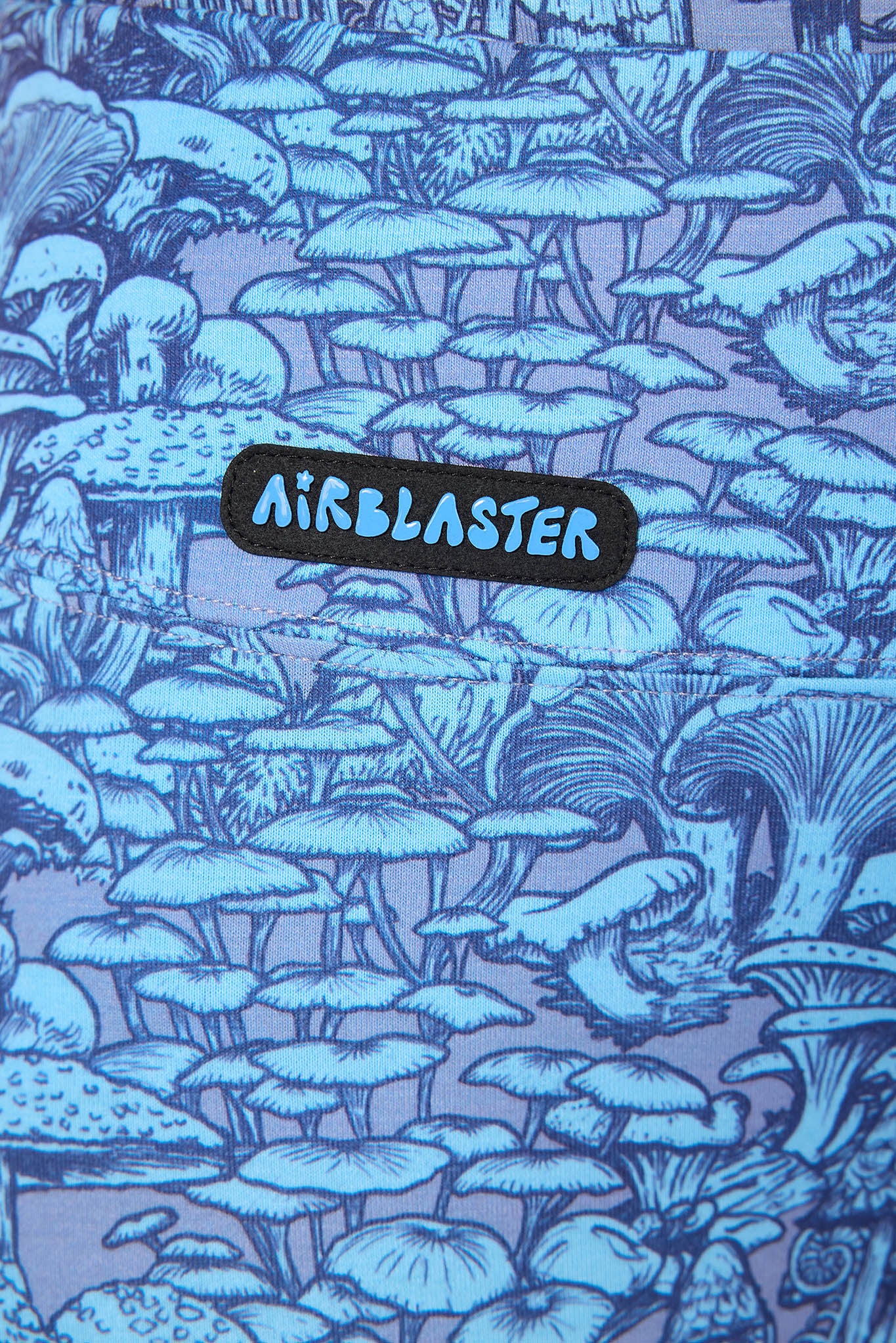 A close-up of a fabric surface featuring an all-over blue and lavender-purple illustrated mushroom pattern, with a black patch reading AIRBLASTER